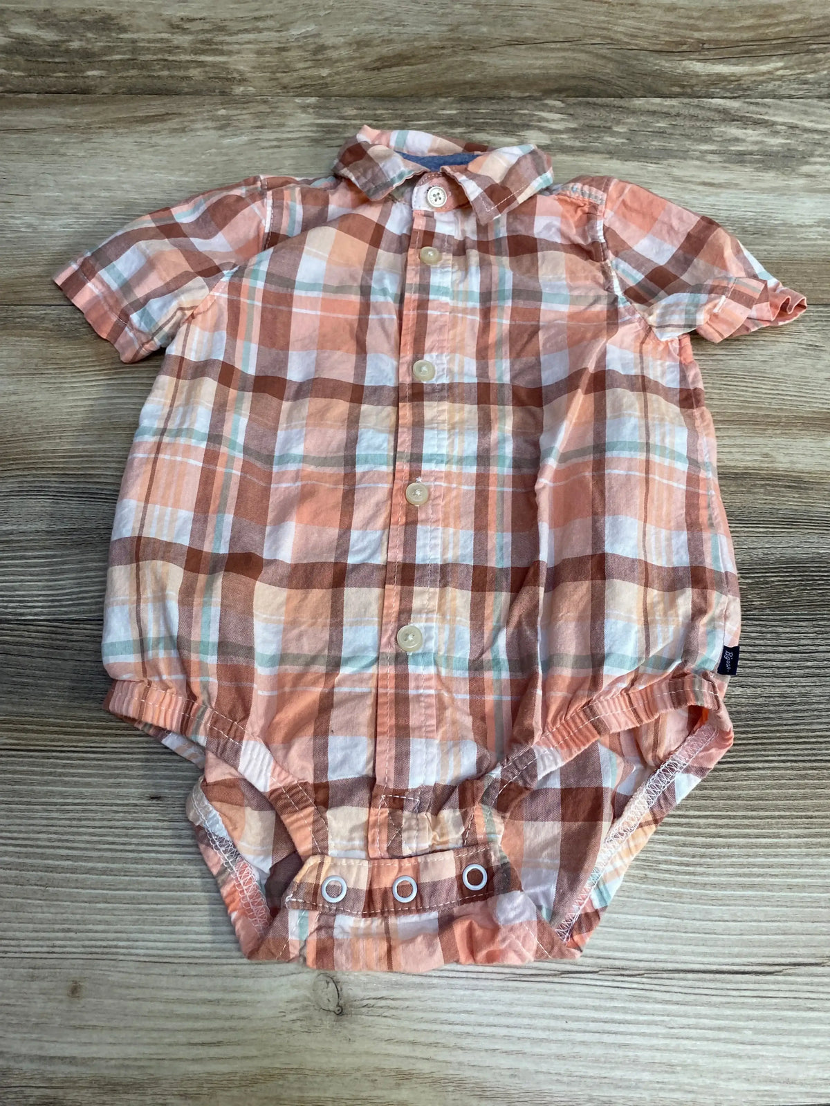 Flat lay of Oshkosh plaid bodysuit in peach orange showing collar, buttons and three snap crotch on wooden background