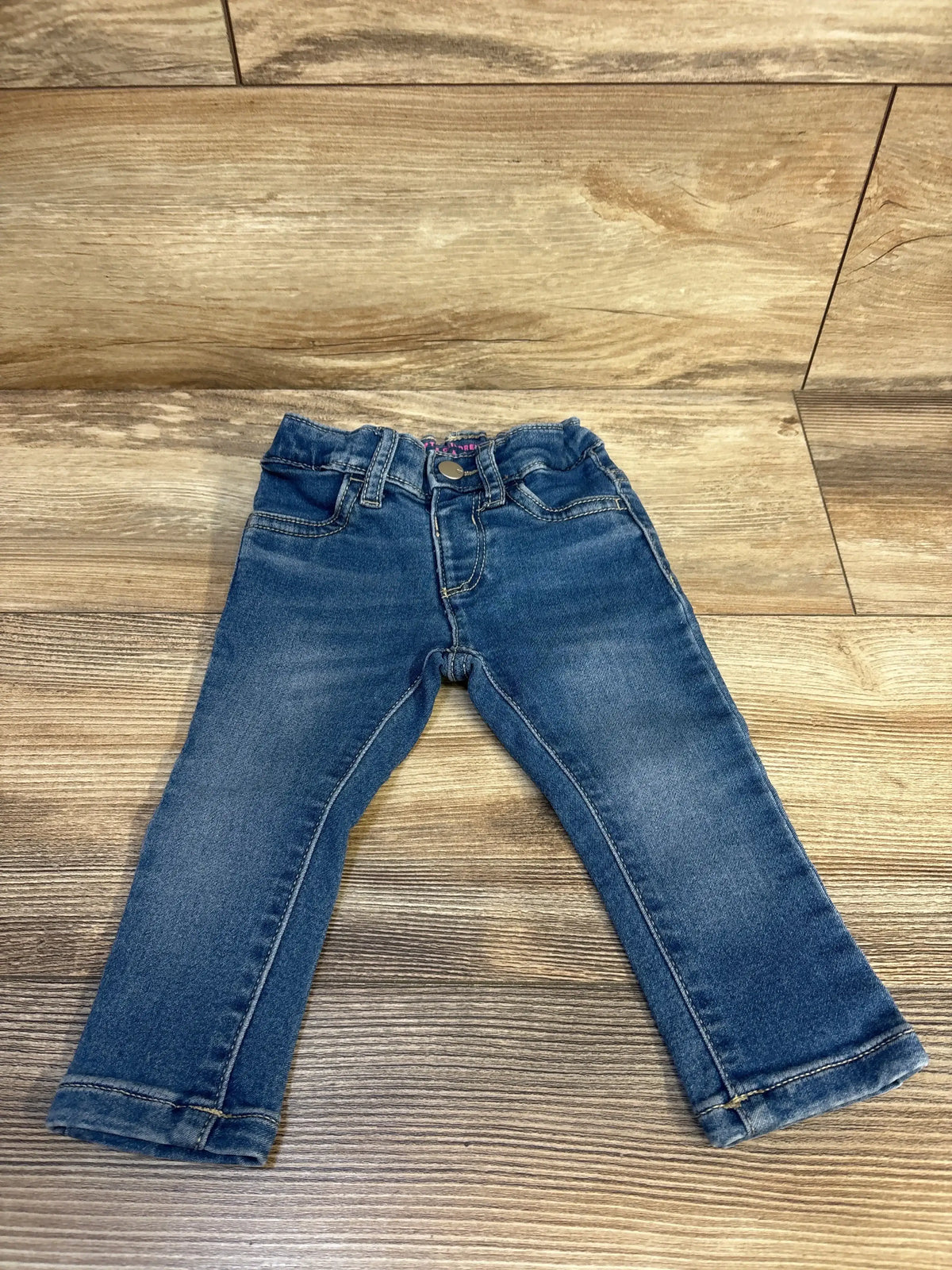Flat lay of Children's Place blue toddler legging jeans size 12 to 18 months showcasing elasticized waistband, faux front pockets and cuffed hems on wood floor