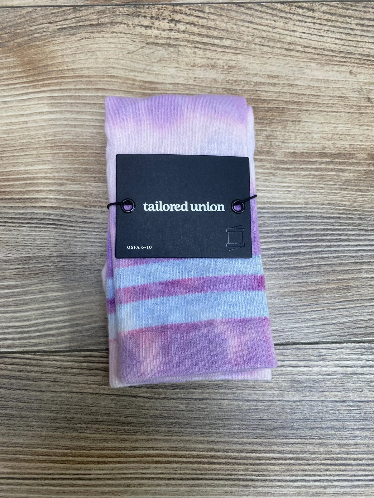 Top view of folded purple-pink socks with pale blue stripes and black Tailored Union tag on wood background