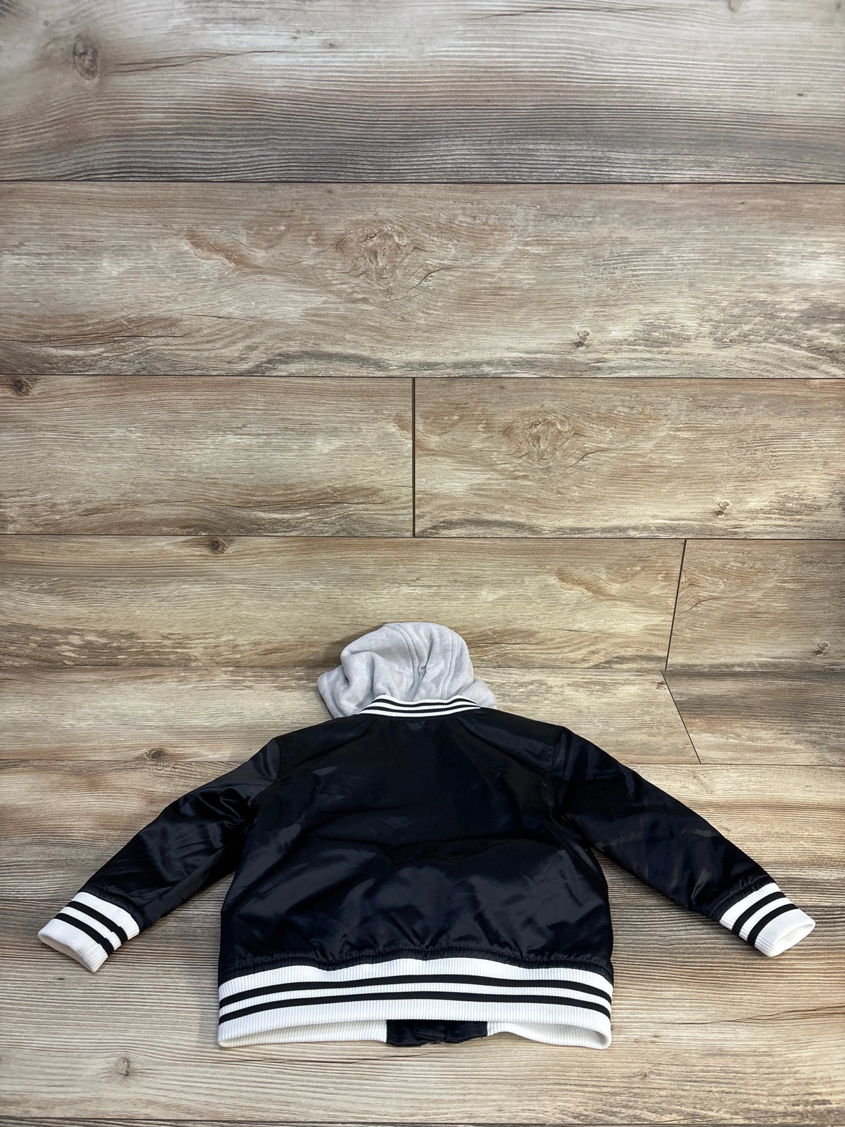 Black kids varsity jacket with white stripes, gray hood, laid flat on wood floor