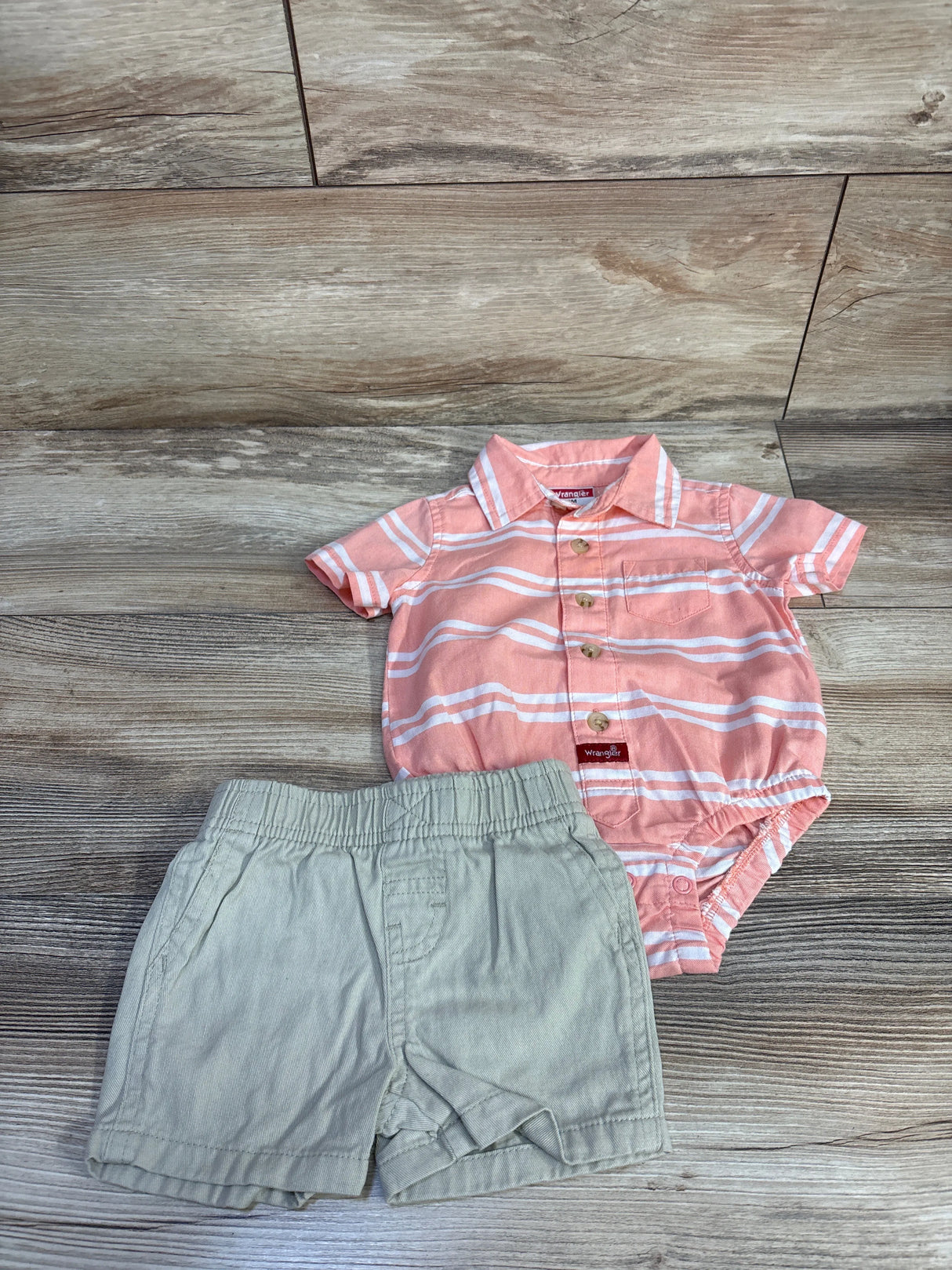 Flat lay of Wrangler pink striped polo bodysuit with khaki shorts, showing front buttons and snaps on wood background