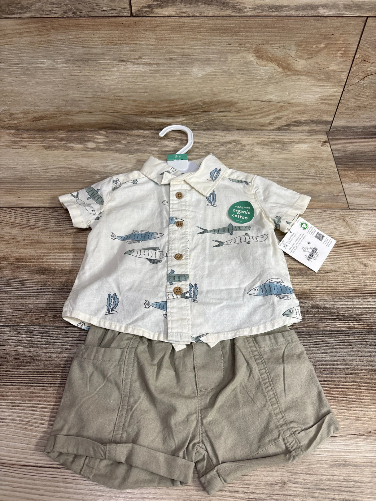Flat lay of 2-piece baby set: fish-print button shirt and khaki shorts, showing organic cotton sticker on wood floor