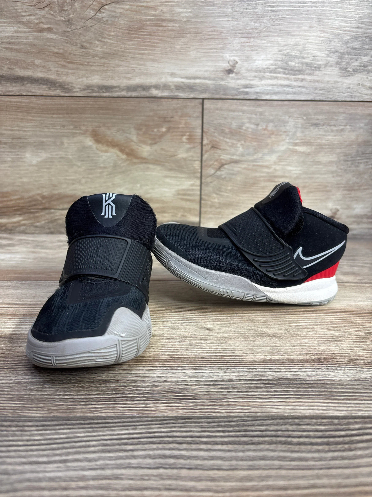 Side view of Nike Kyrie kids athletic sneakers showcasing black upper white sole and velcro strap closure on wood plank floor