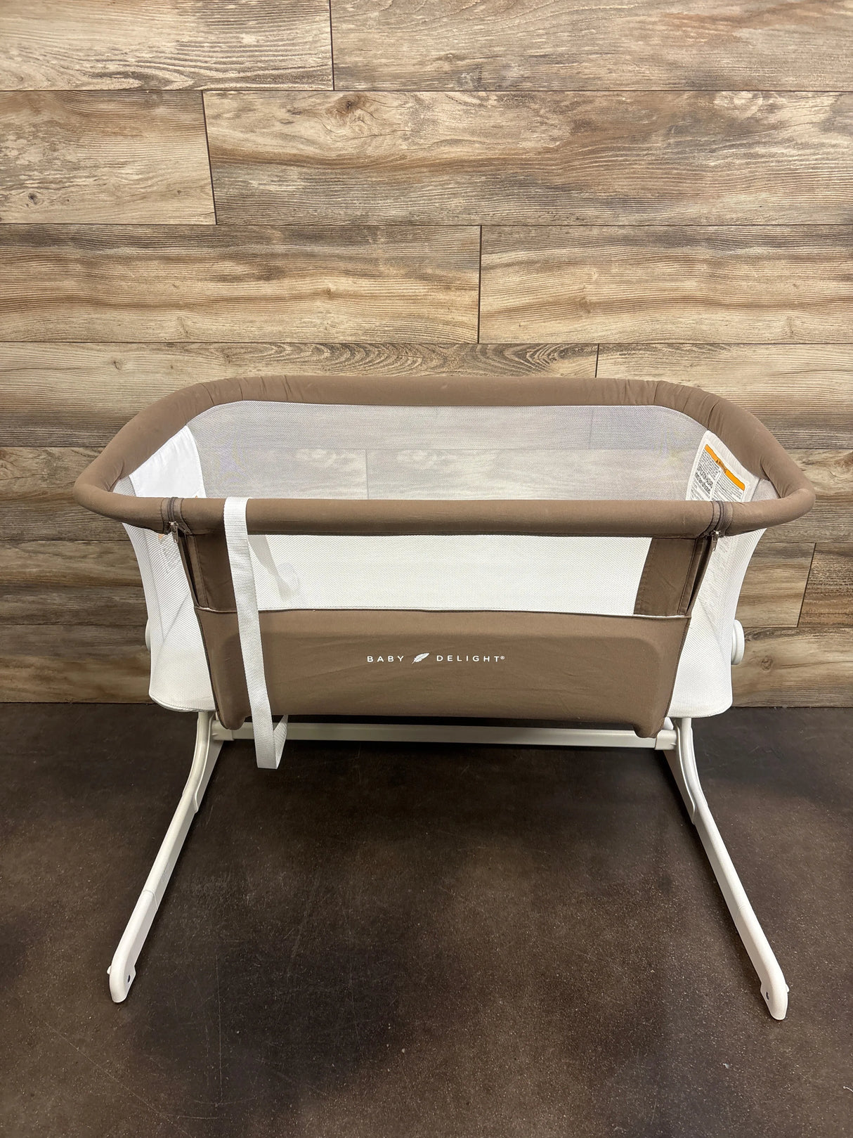 Front view of Baby Delight Beside Me Dreamer bedside bassinet in mocha with mesh sides and white legs on floor and wood wall