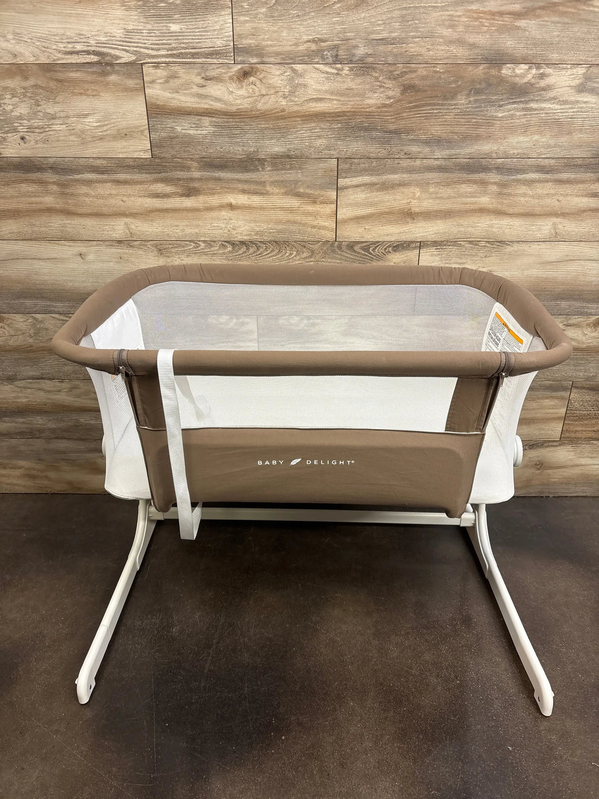 Front view of Baby Delight Beside Me Dreamer bedside bassinet in mocha with mesh sides and white legs on floor and wood wall