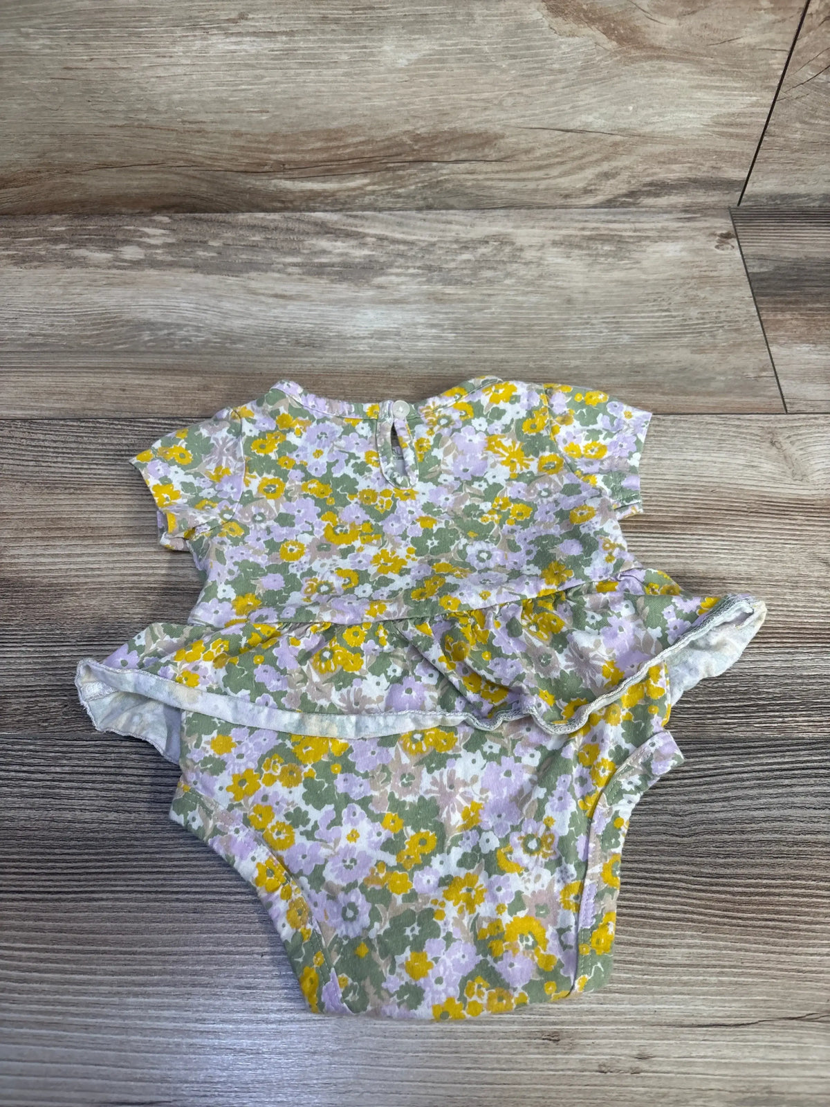 Top-down view of green floral baby bodysuit dress showcasing ruffled peplum, short sleeves, and snap crotch on wood floor