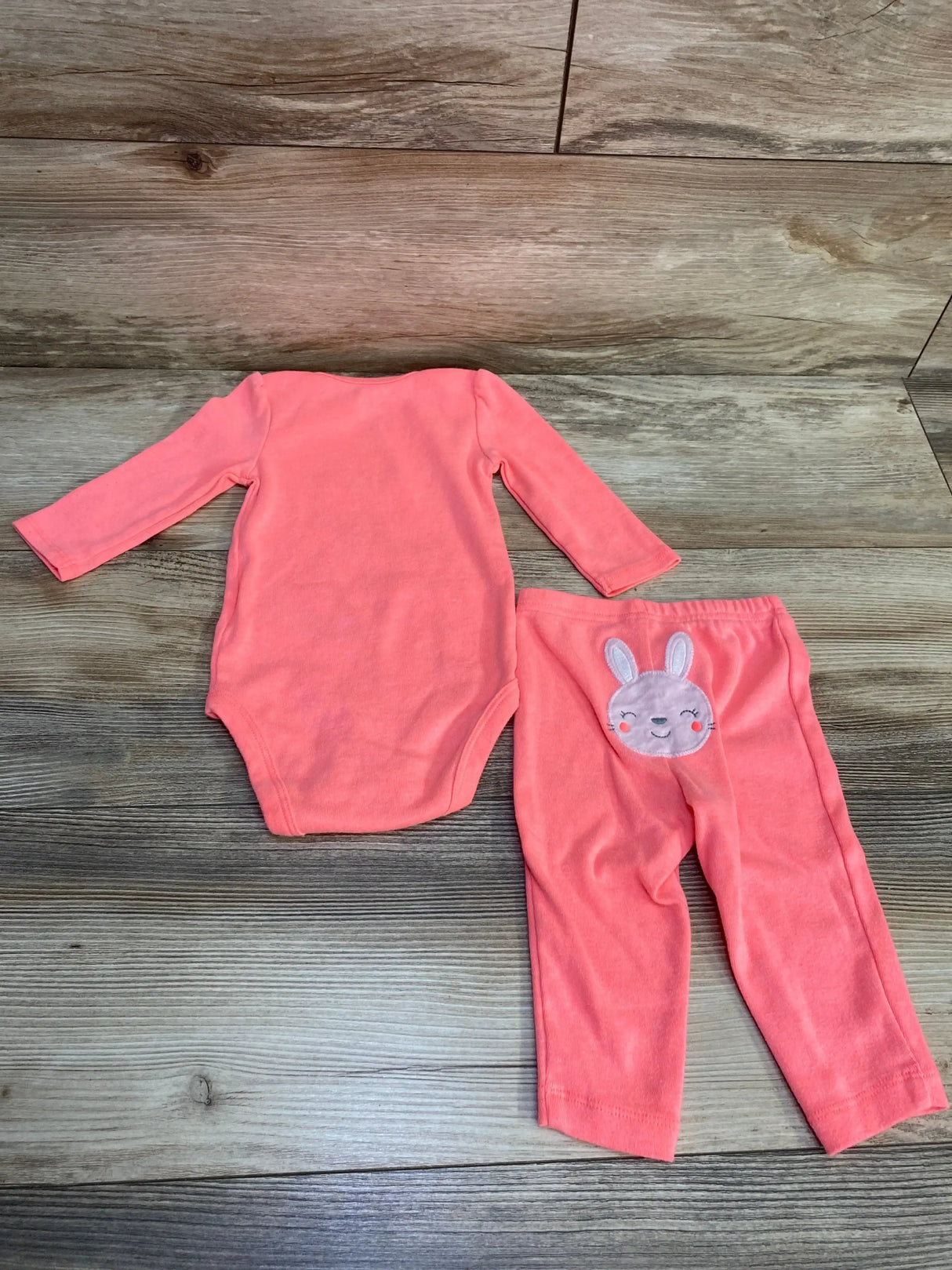 Flat lay of Simple Joys 2pc bunny bodysuit and pants in pink showcasing long-sleeve bodysuit and bunny applique on pants with wood plank floor