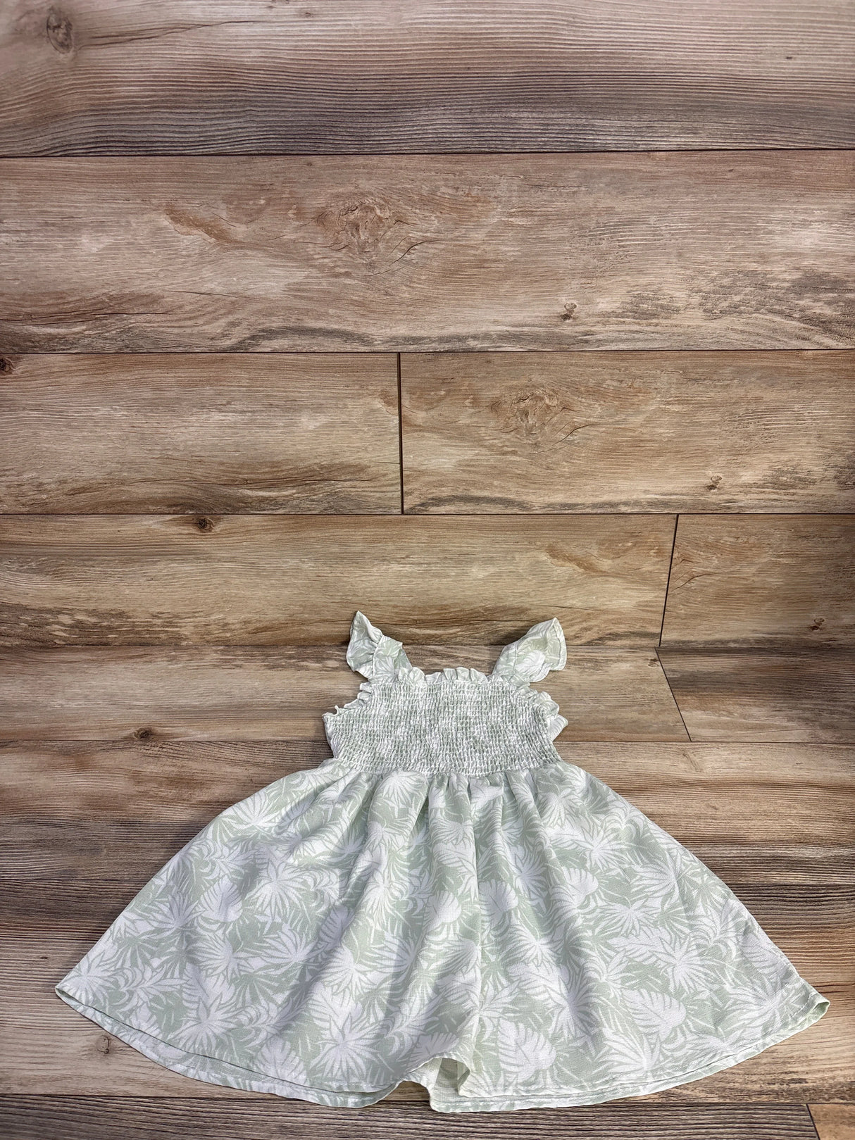 Flat lay of pale green Little Lass smocked girls dress with ruffle shoulder straps and white leaf print on wood background