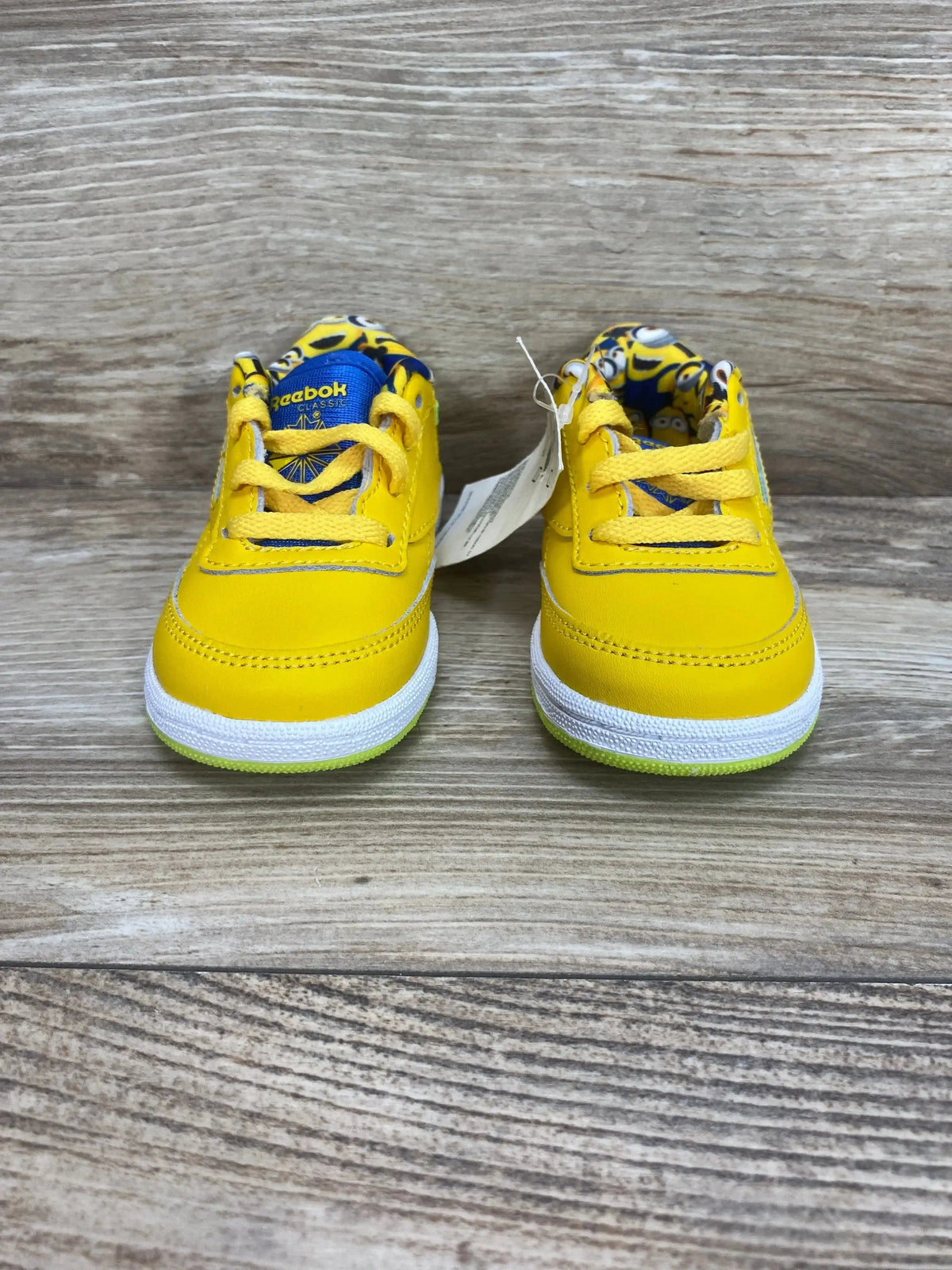Front view of toddler Club C85 Minions sneakers with bright yellow upper and Minions-print lining on wood background