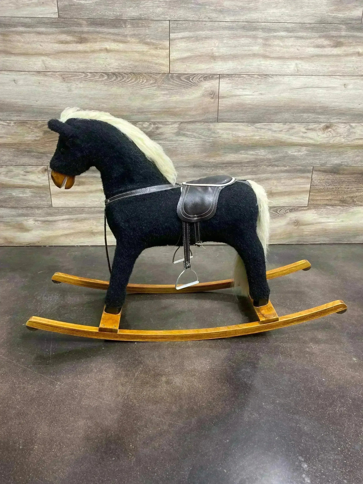 Side view of vintage black plush rocking horse showcasing cream mane and tail, leather saddle and wooden rockers on a wood-paneled floor