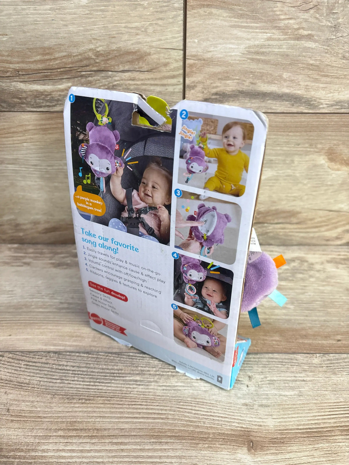 Angled view of Fisher-Price Sing & Go purple monkey stroller toy showing plush face, colored ribbon tags, and green clip ring on wood background