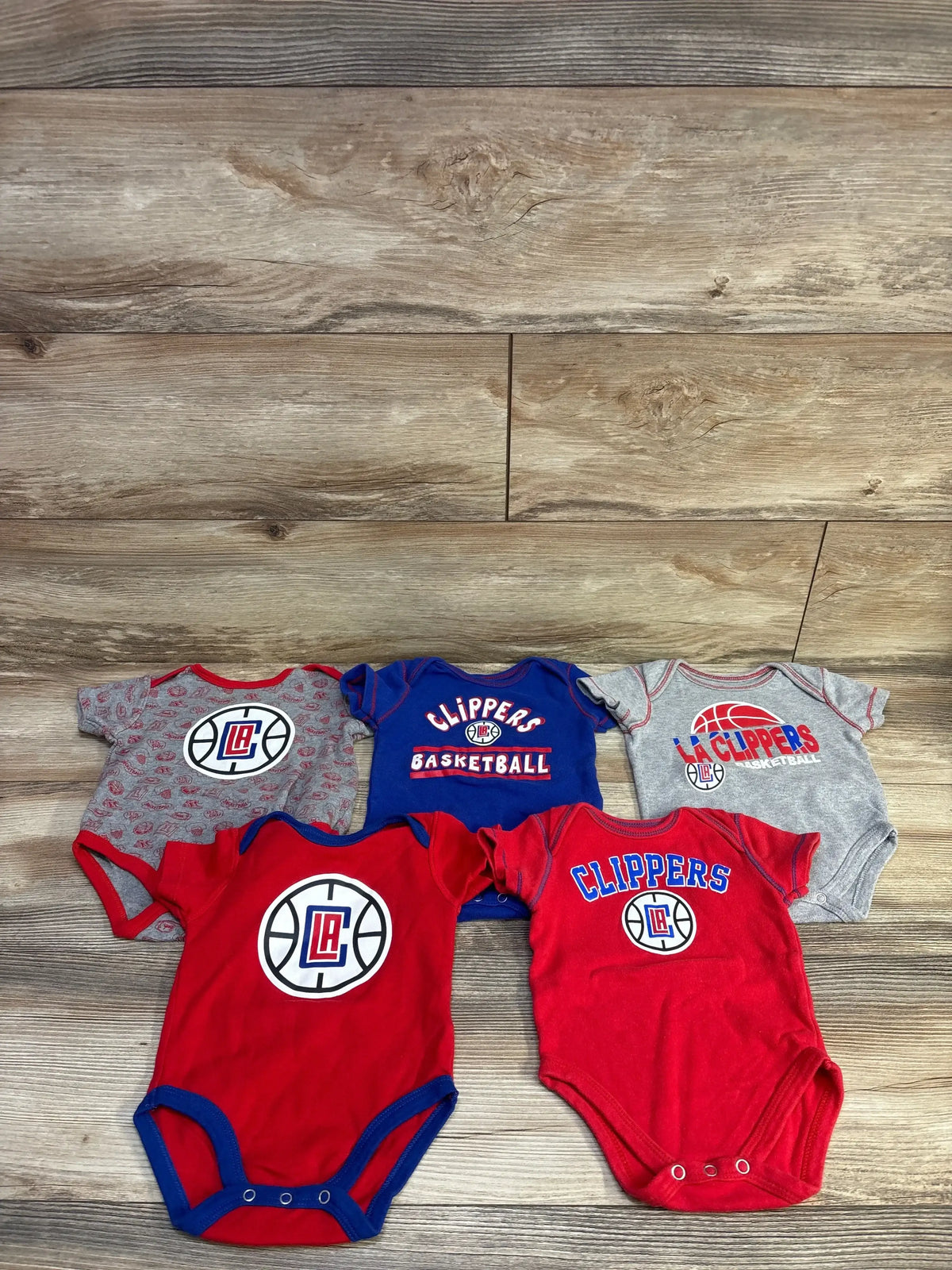 Top view of five NBA Clippers bodysuits showcasing screen-printed basketball designs and three-snap closures on wood