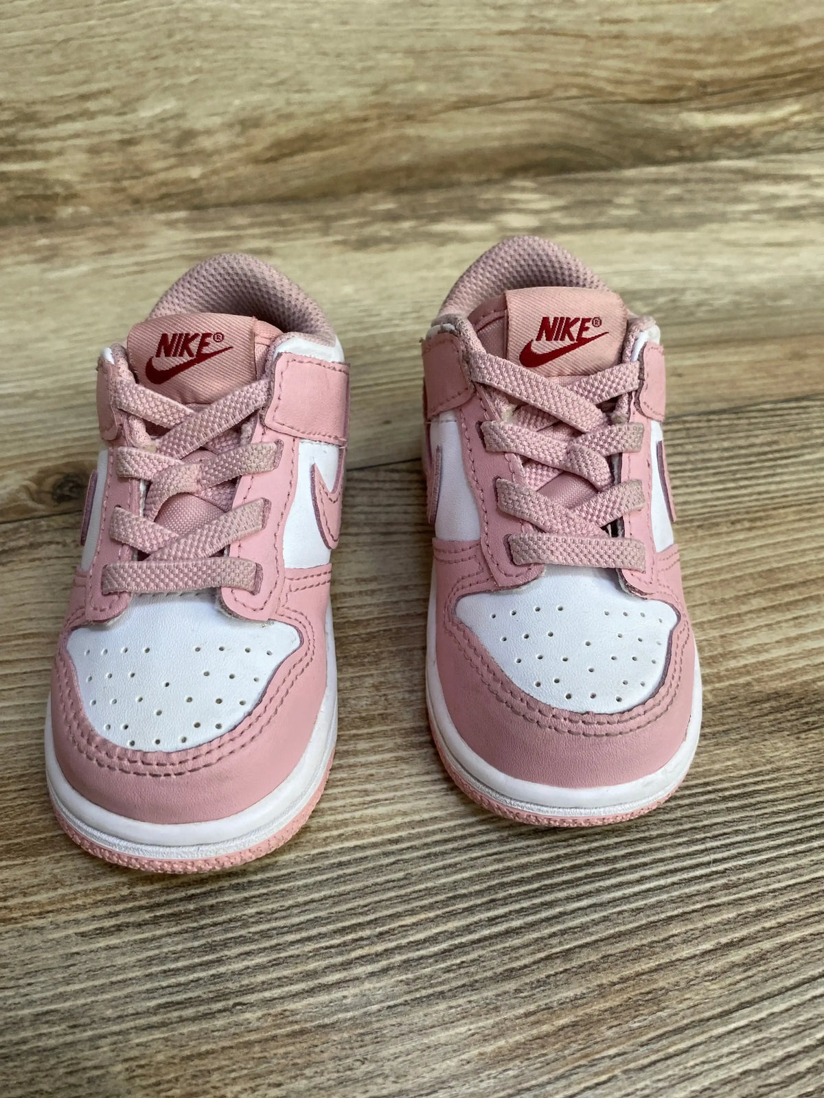 Front view of toddler Nike Dunk Low Pink Glaze sneakers showing pink and white leather panels, laces, and red Nike logo on the tongue on a wood floor