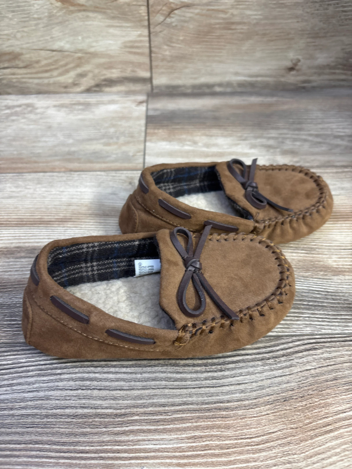 Angled side view of Cat & Jack Lionel toddler moccasin slippers in brown showing stitched toe, bow detail, and fleece lining on wood background