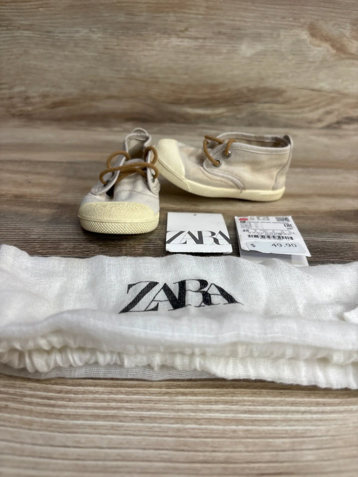 Pair of beige Zara toddler shoes with laces, price tag, and Zara branded cloth bag on wooden surface