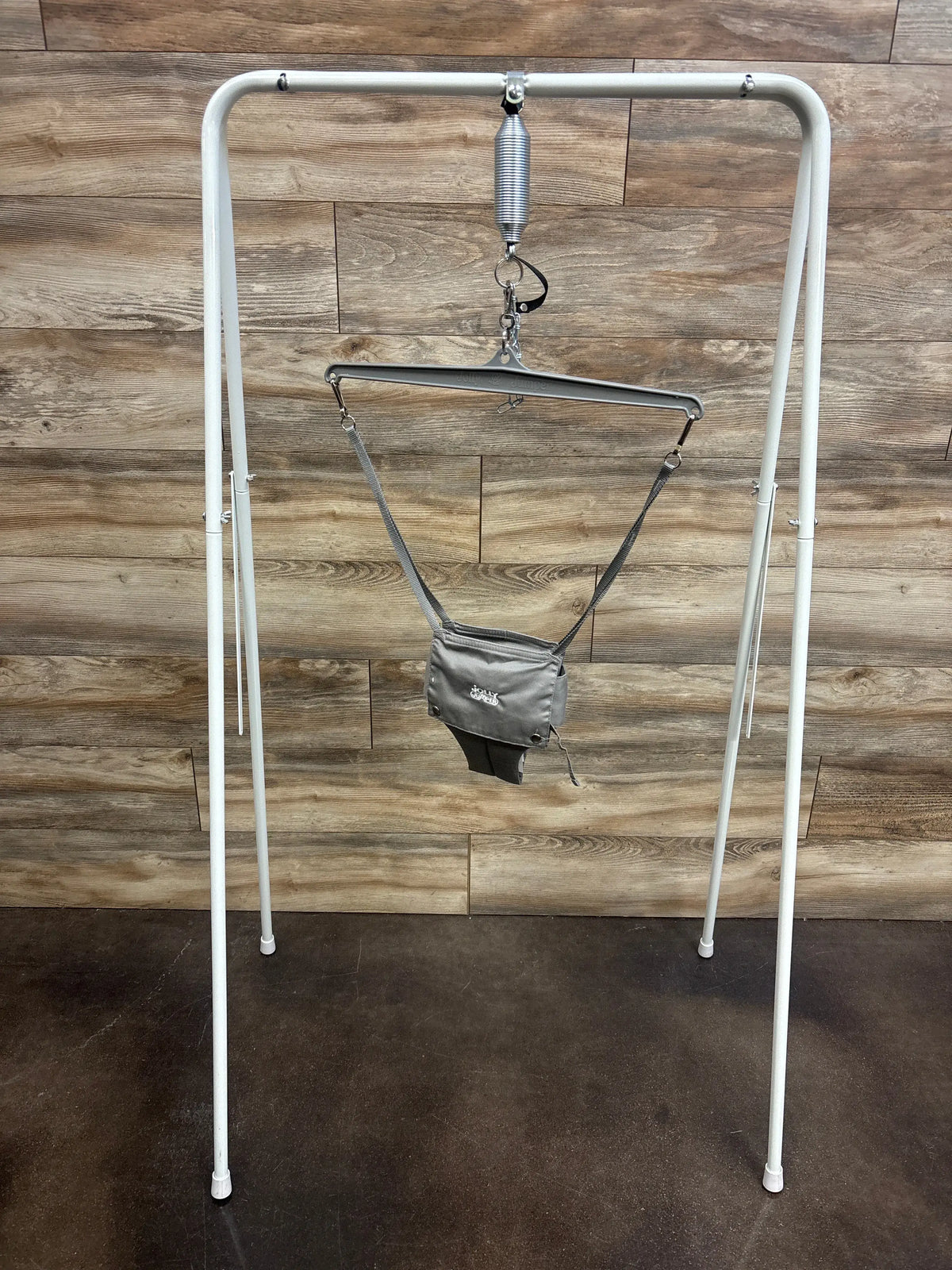 Front view of Jolly Jumper Classic with stand showing grey fabric jumper seat suspended from metal spring and hanger against wood wall