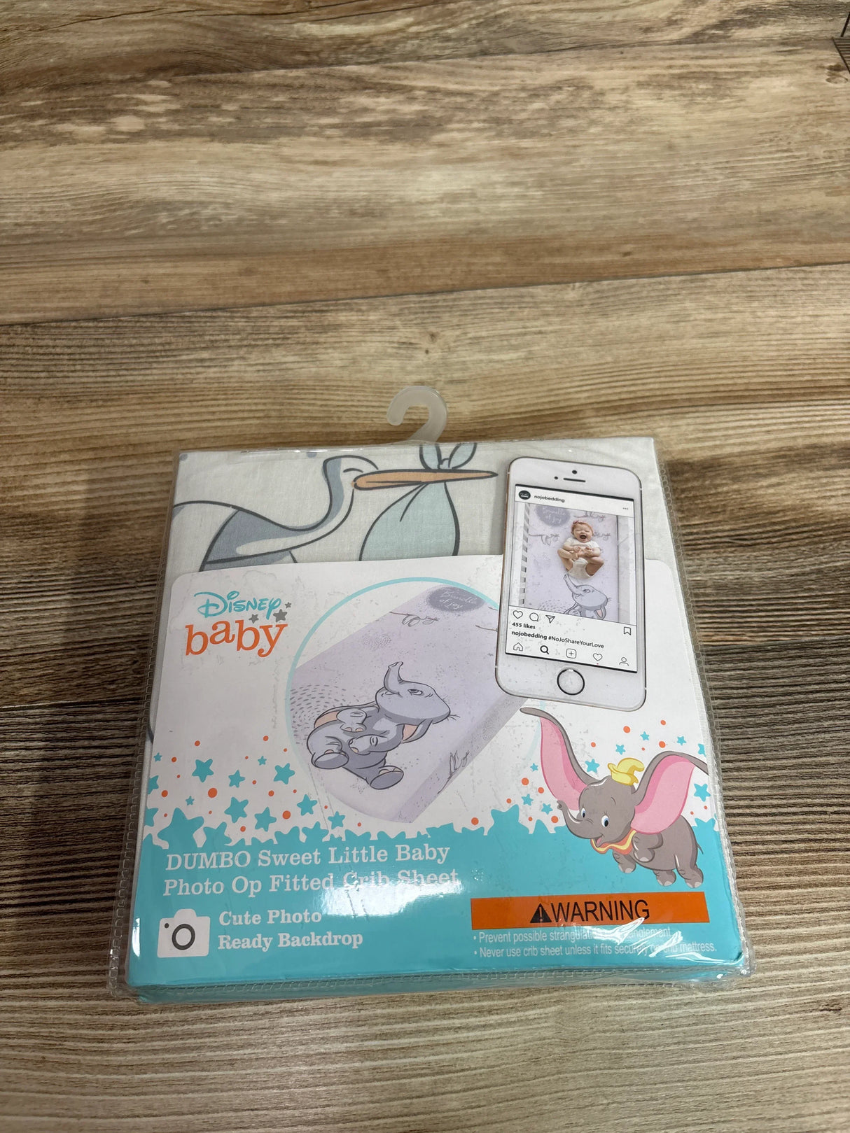 Front view of packaged Disney Baby Dumbo fitted crib sheet showcasing Dumbo print and photo-op backdrop graphic on wooden table