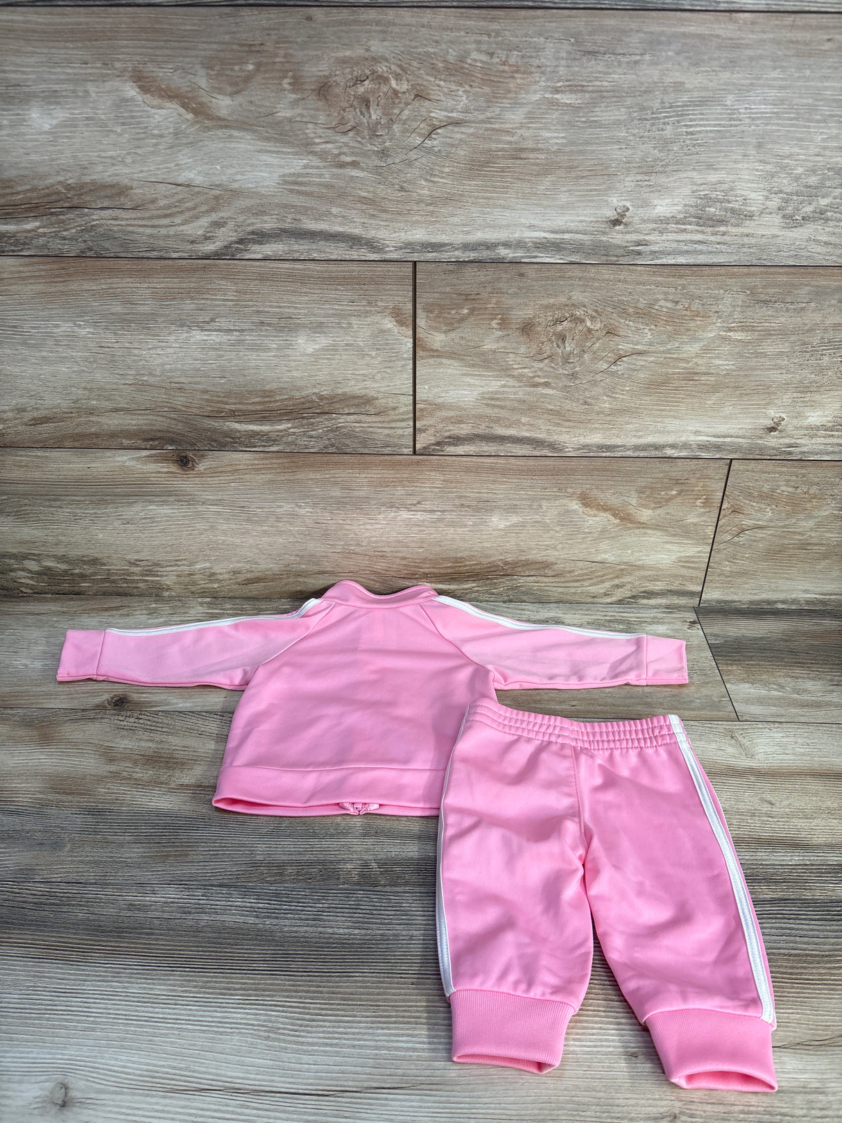 Flat-lay of baby pink Adidas 2-piece tracksuit showing jacket back with white three stripes and matching pants with elastic waist on wood background
