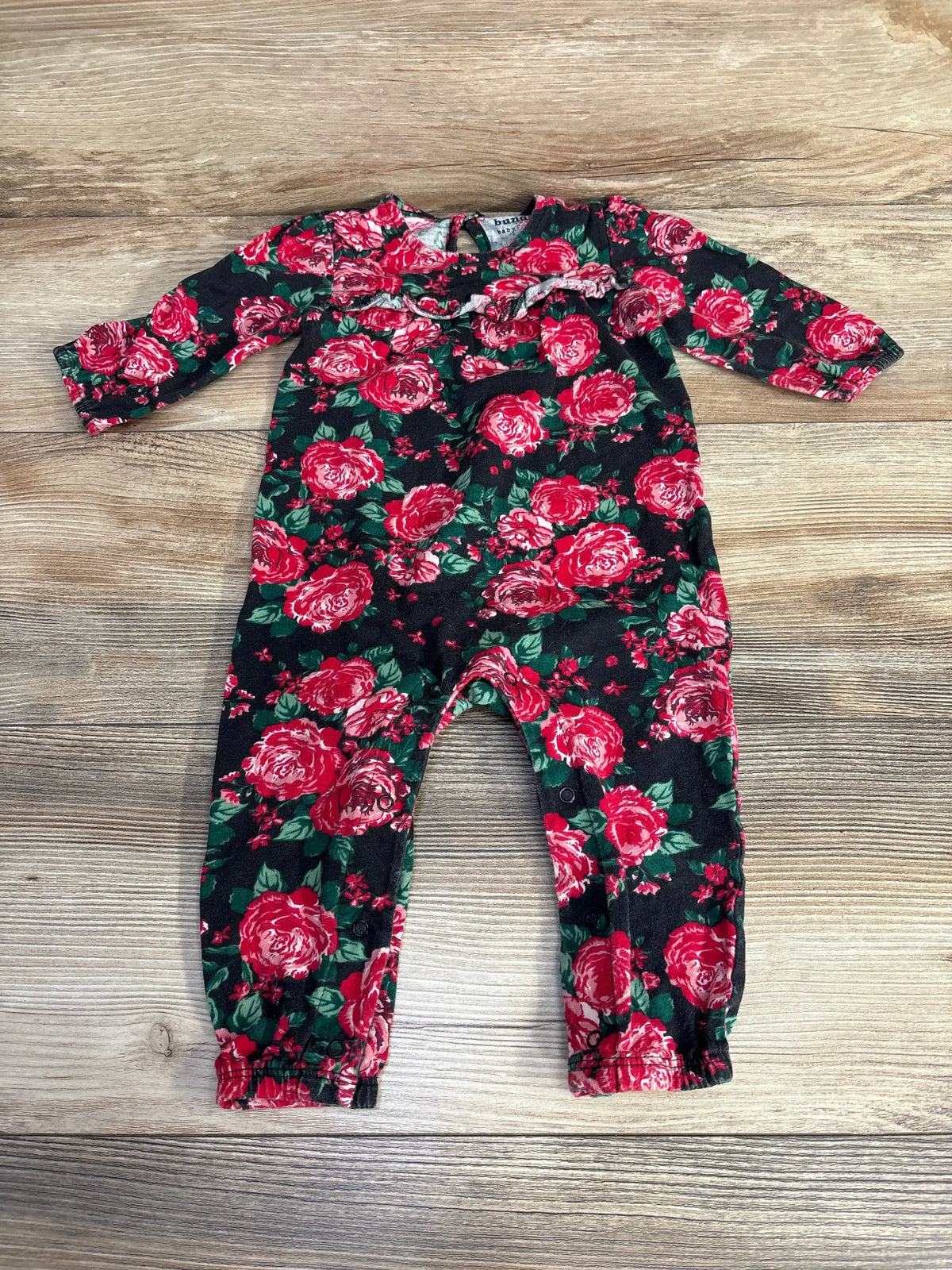 Flat lay of baby floral coverall showcasing red rose print, chest ruffle and snap leg closures with wood background