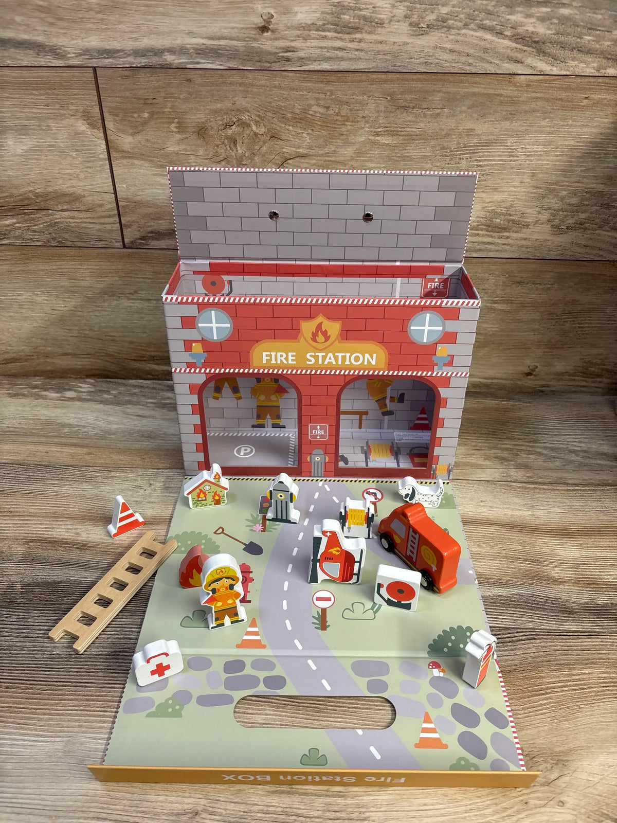 Angled view of Mud Pie wooden fire station playset 14pc showing fold-out mat, toy truck, figures, ladder on wood tabletop