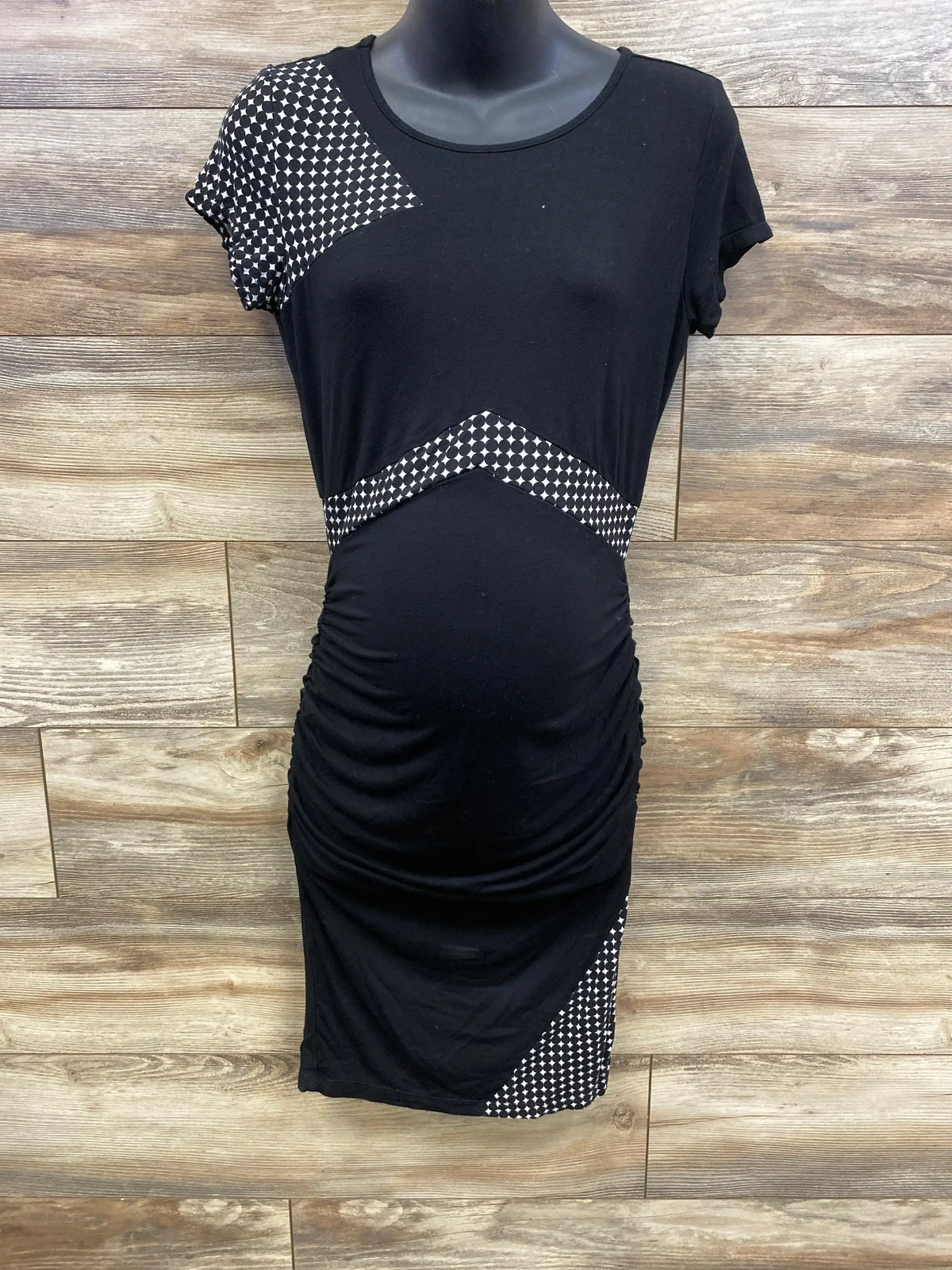 Front view of black Kimi + Kai bodycon maternity dress on mannequin, ruched sides and chevron contrast panels, wood backdrop