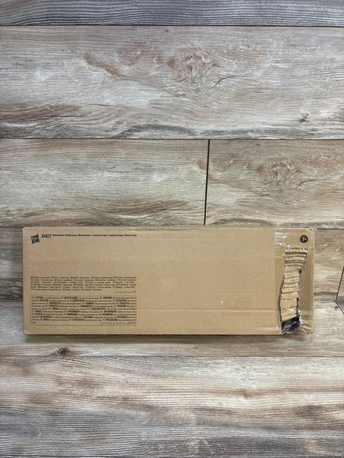 Front view of Nerf Super Soaker Barrage shipping box showing unopened brown cardboard with torn corner on wood floor