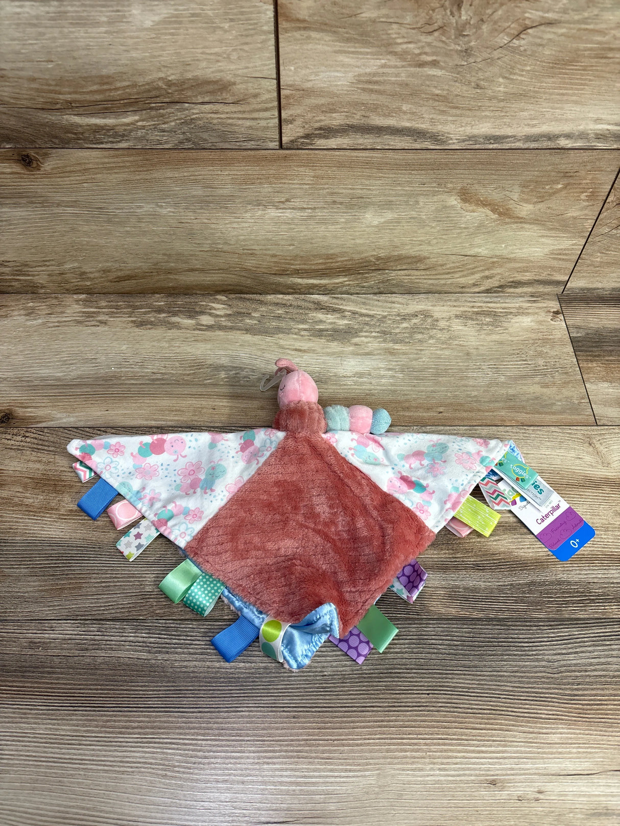 Soft plush pink and white caterpillar baby blanket with colorful ribbon tags on wood floor