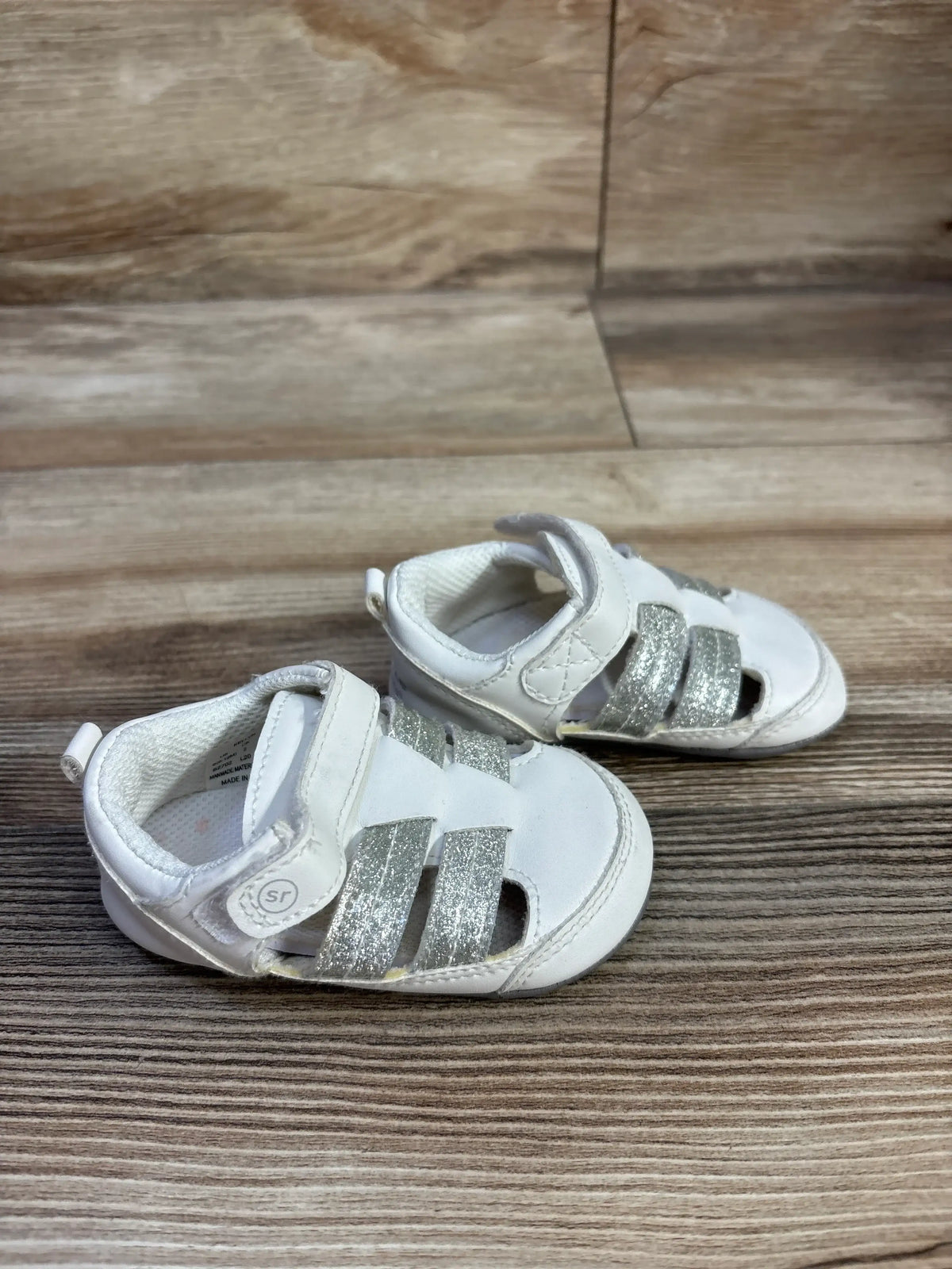 Angled view of Surprize by Stride Rite white fisherman sandals, size 3C, with silver glitter straps and adjustable closures on wood-look flooring