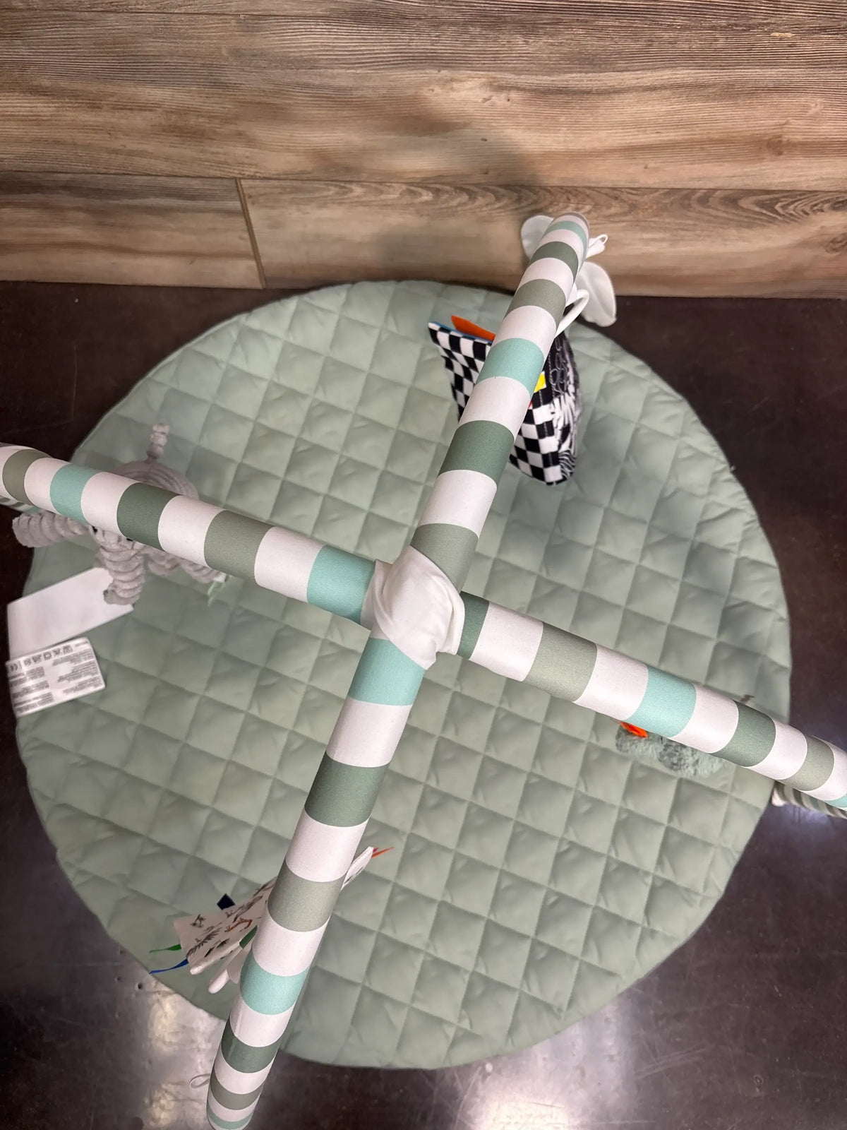 Top view of baby play gym activity mat in sage green showing quilted padding, striped arches, and hanging toys on a wood floor