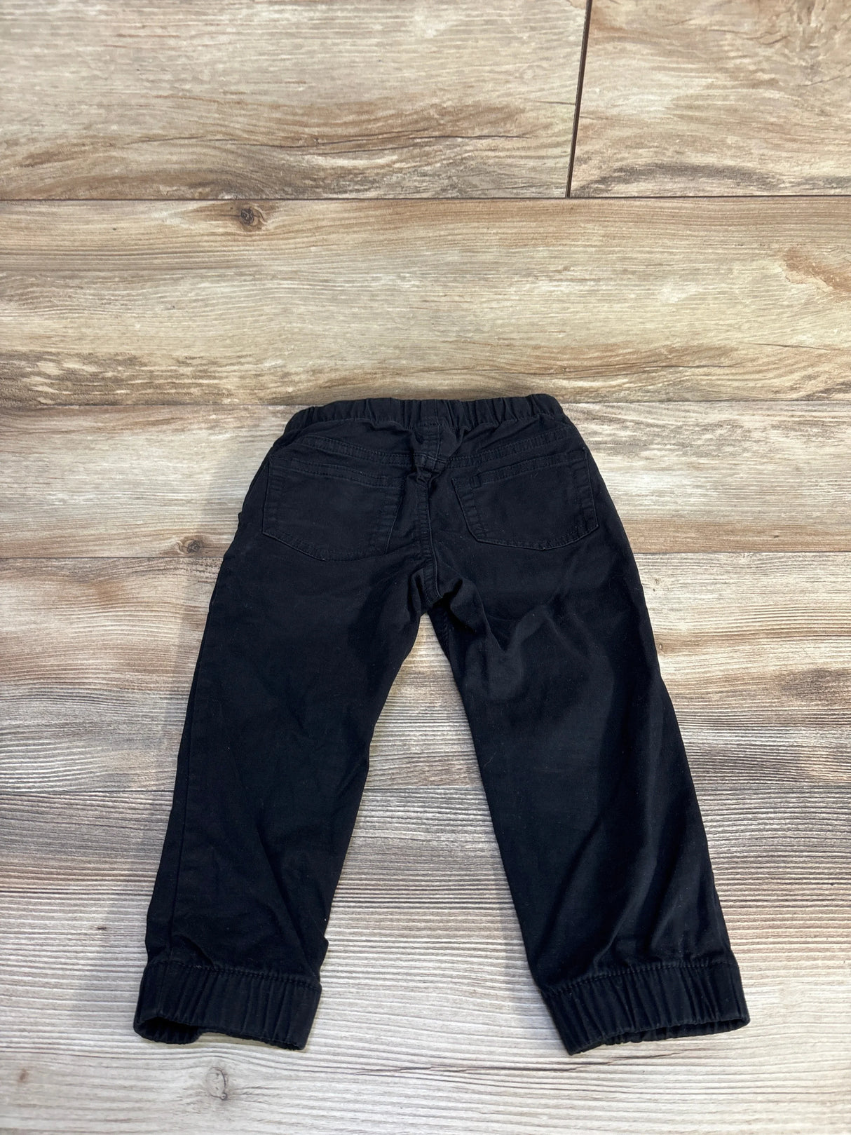 Back view of black toddler drawstring joggers (2T) showing elastic waistband, back pockets, and ribbed cuffs on wood background