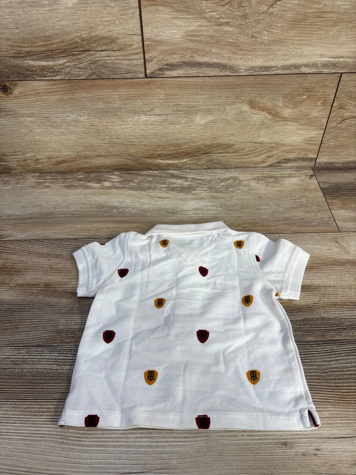 Back view of white Tommy Hilfiger polo shirt size 12m showcasing embroidered red and yellow shield motifs on wood floor