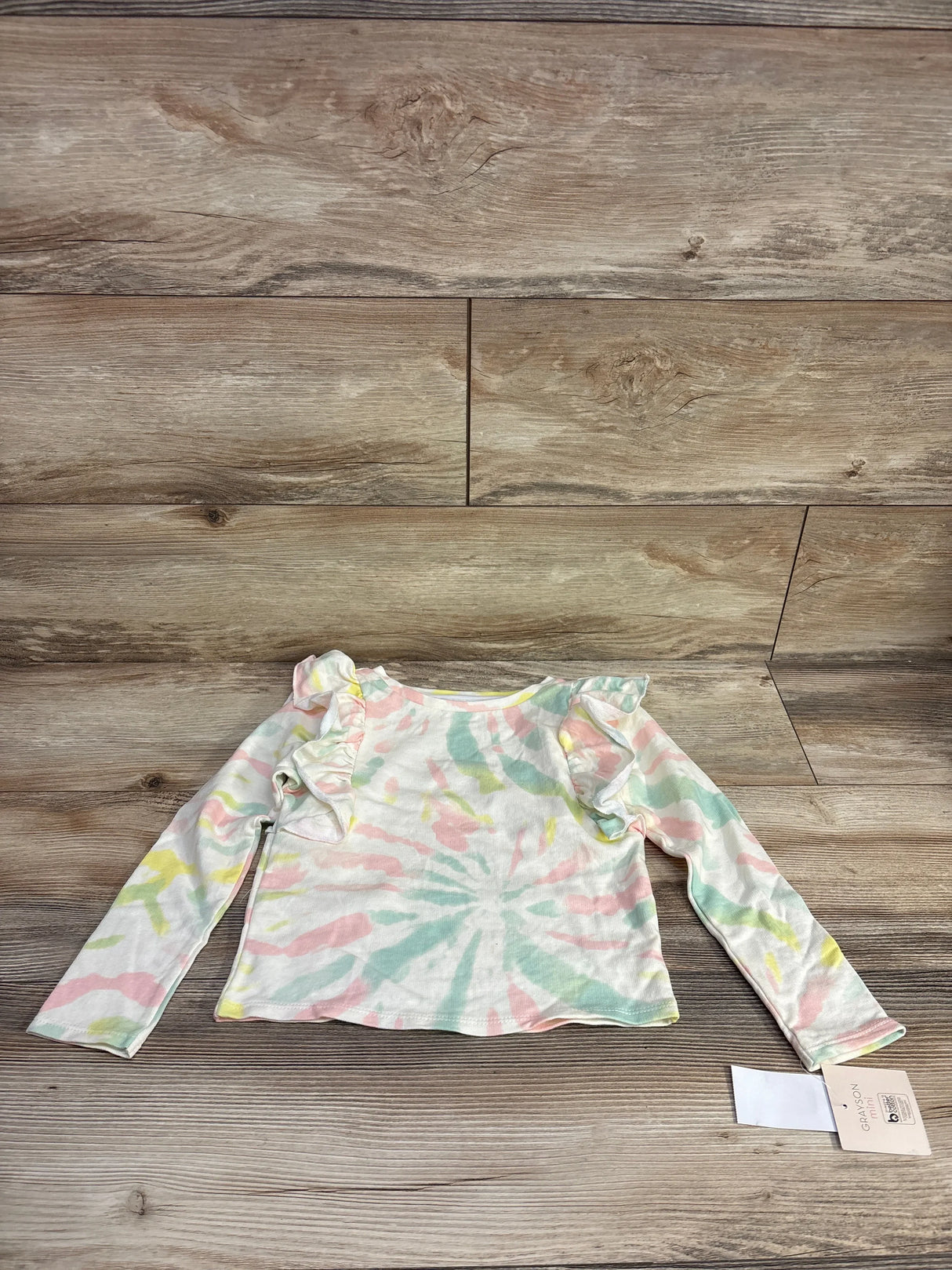 Flat lay of NEW Grayson Mini Tie-Dye Ruffle Top in pastel pink green and yellow swirl with shoulder ruffles and hangtag on wood plank floor