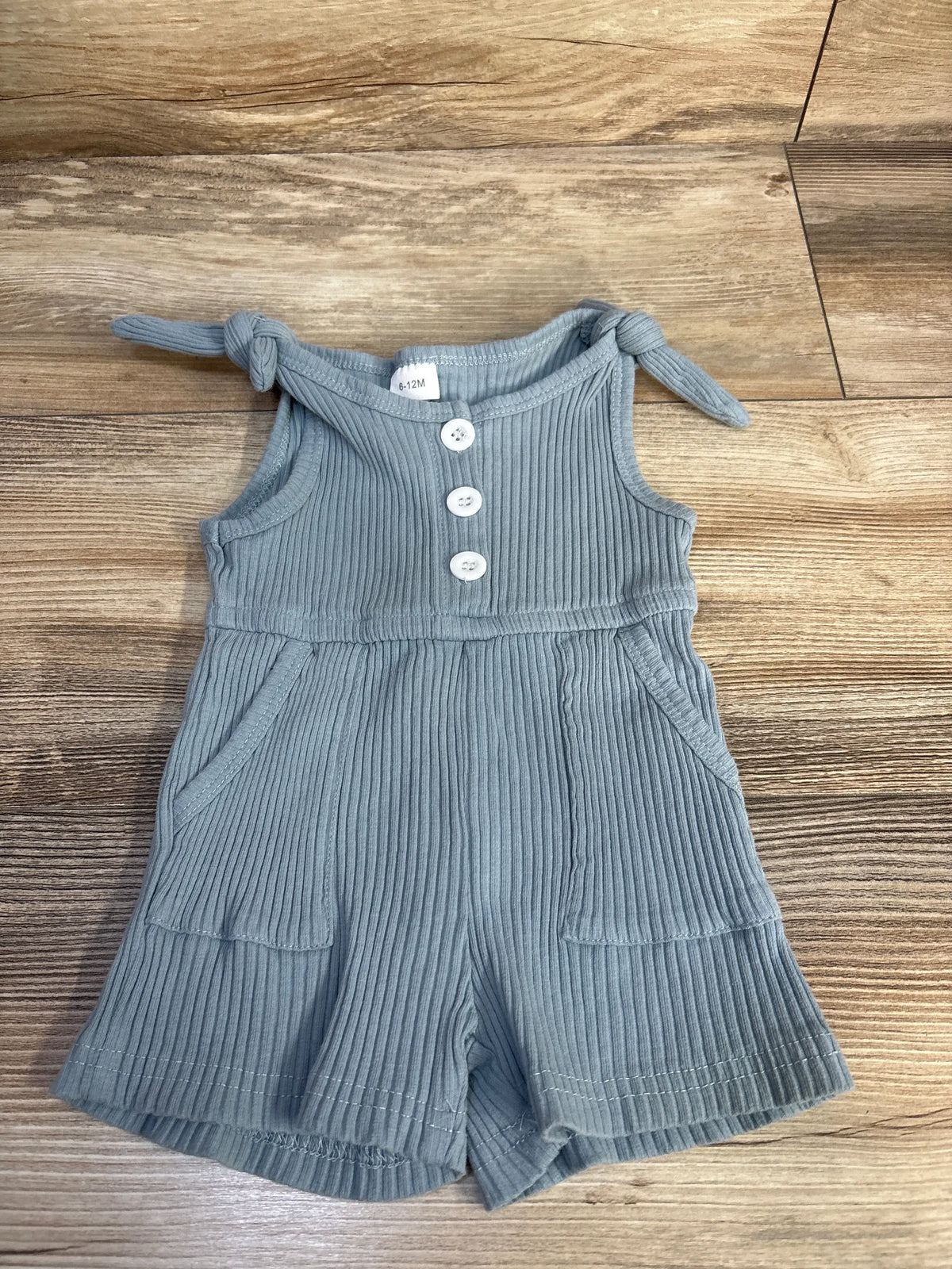 Top view of blue ribbed tank shortie romper showcasing tie shoulder straps, three buttons, and pockets on a wood background