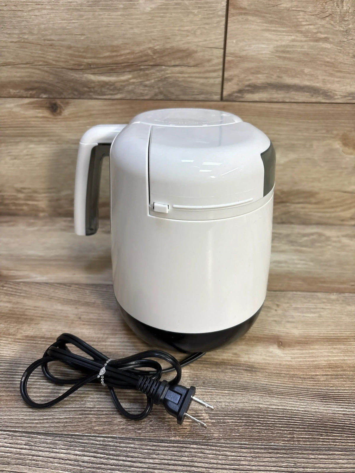 Front view of Baby Brezza One Step Baby Food Maker Deluxe showcasing white housing, black base, and attached power cord on a wood surface