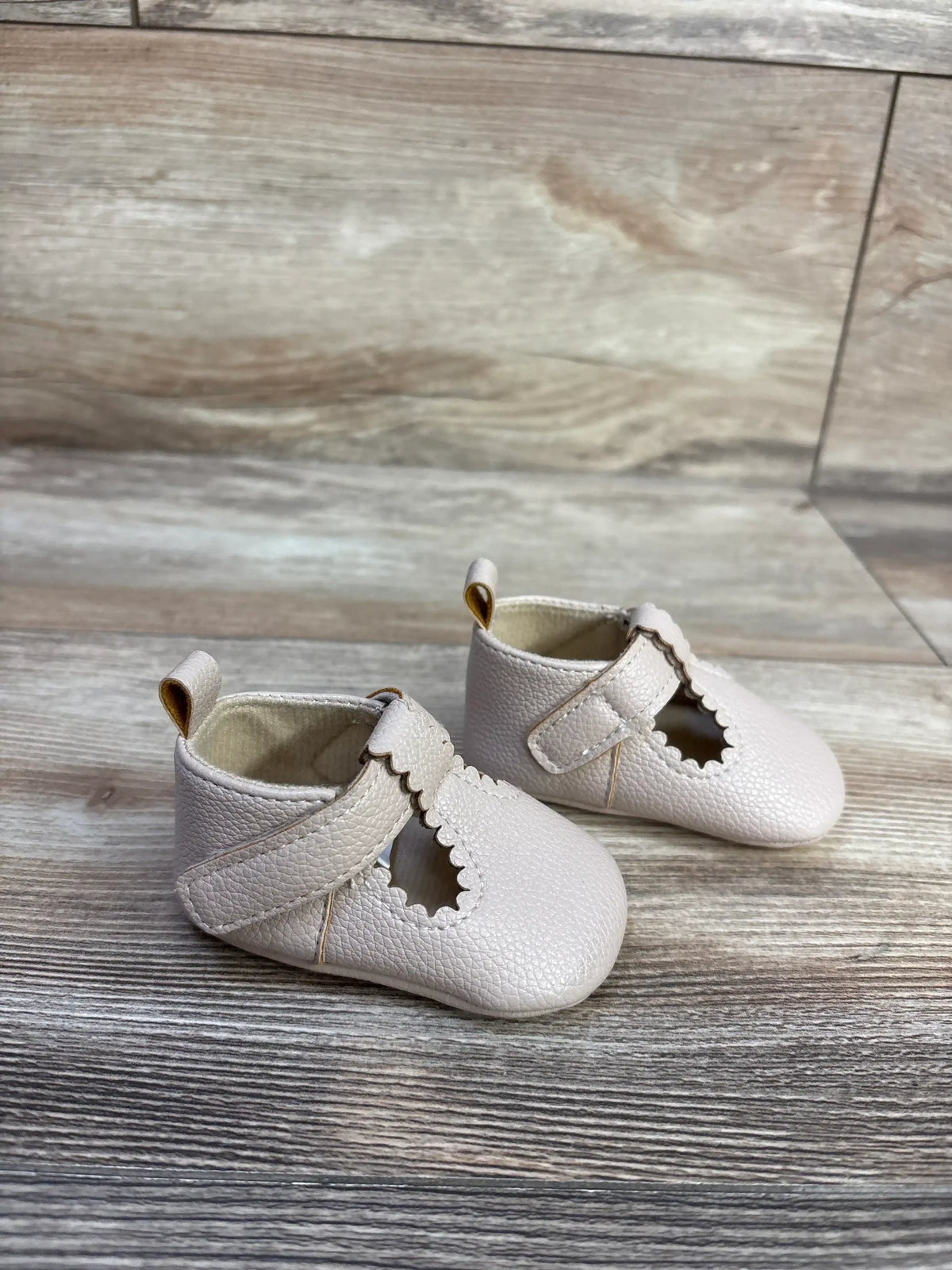 Three-quarter view of beige soft-sole baby shoes with scalloped T-strap and pebbled leather on light wood background