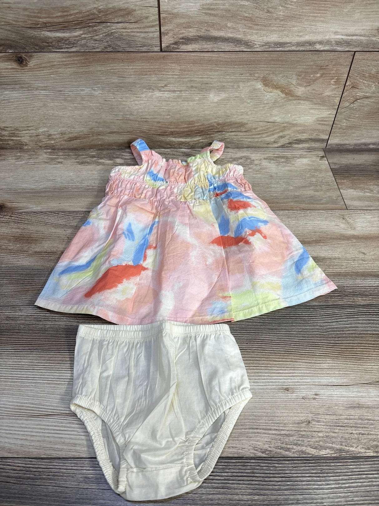 Flat lay of pink multicolor baby dress showing smocked ruffle bodice with matching white bloomers on wooden floor