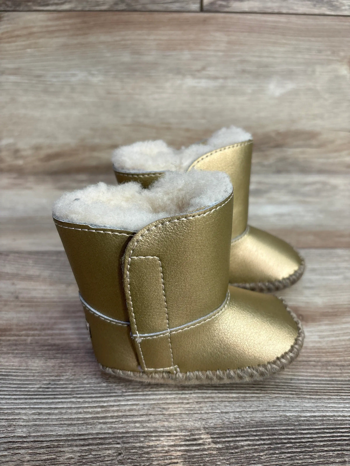 Three-quarter view of baby UGG Cassie boots showcasing metallic gold finish, white shearling lining, and stitched sole on rustic wooden surface