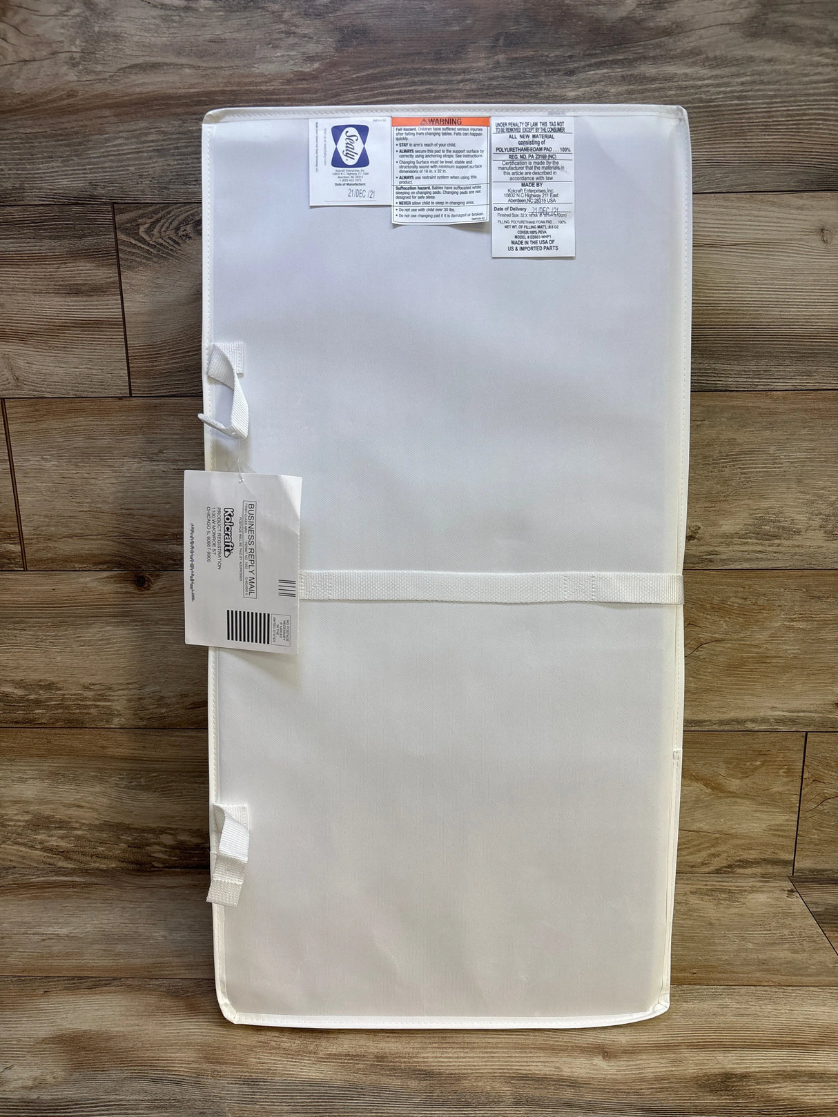 Top-down view of Kolcraft Sealy Soybean Comfort Contoured Changing Pad showcasing contoured white wipeable surface and attached safety straps with wood floor background