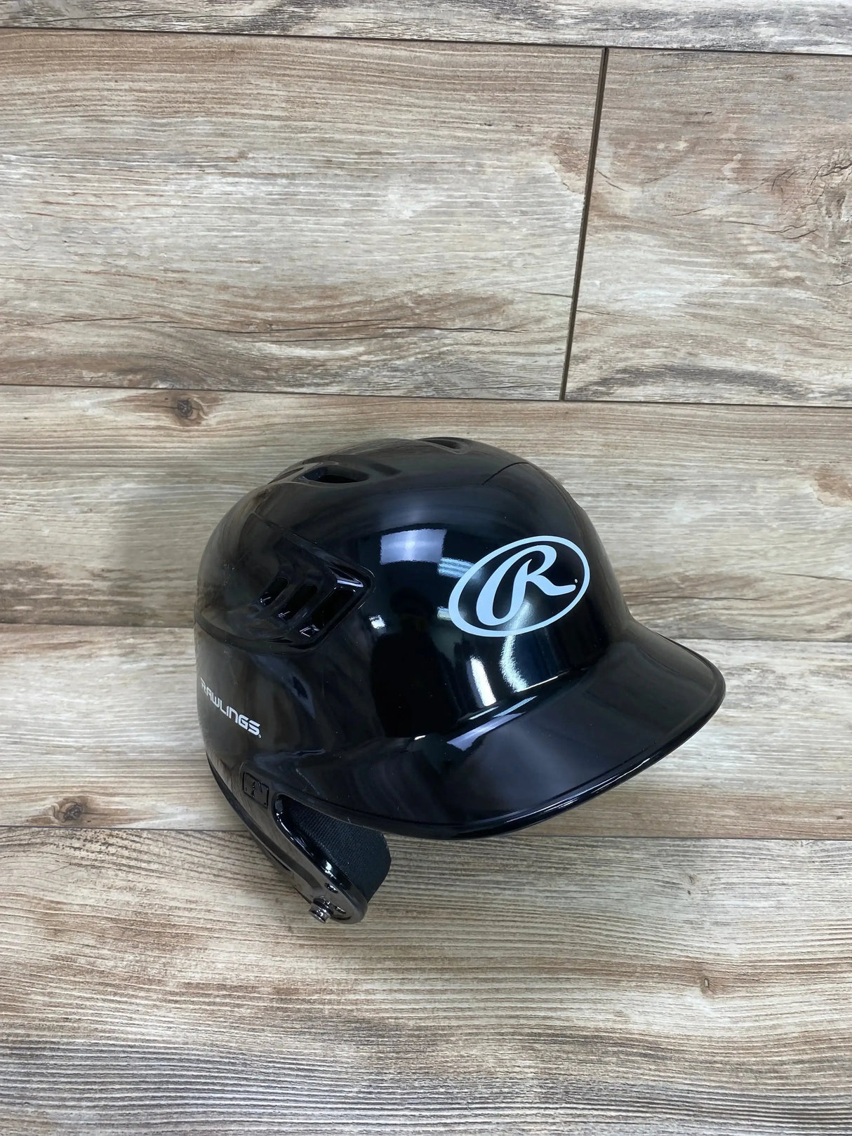 Three-quarter view of Rawlings black batting helmet showing glossy finish, right ear flap, and white Rawlings logo on wood plank background