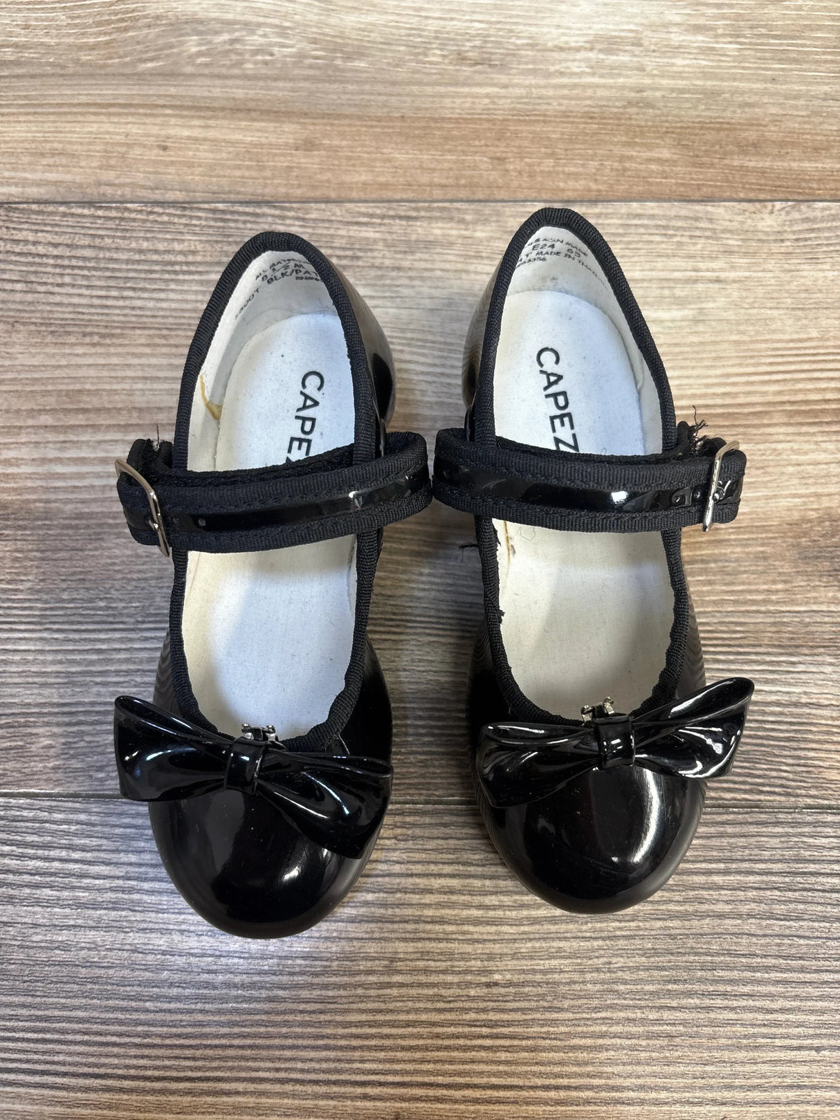 Black patent leather girls' Mary Jane shoes with bows and buckle straps on wood floor