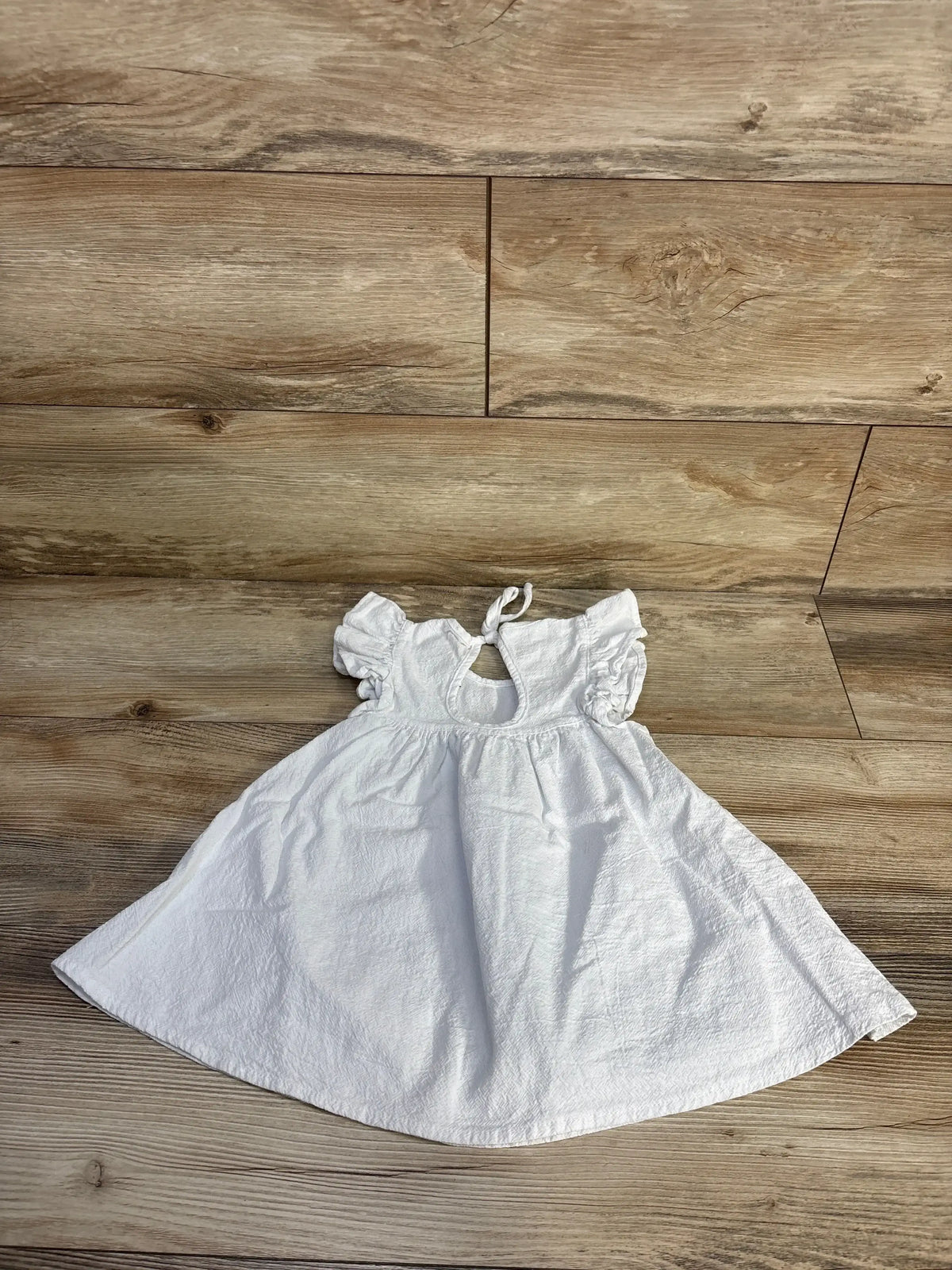 Flat-lay of white flutter-sleeve textured toddler dress (18m) with keyhole back tie and gathered skirt on wood floor