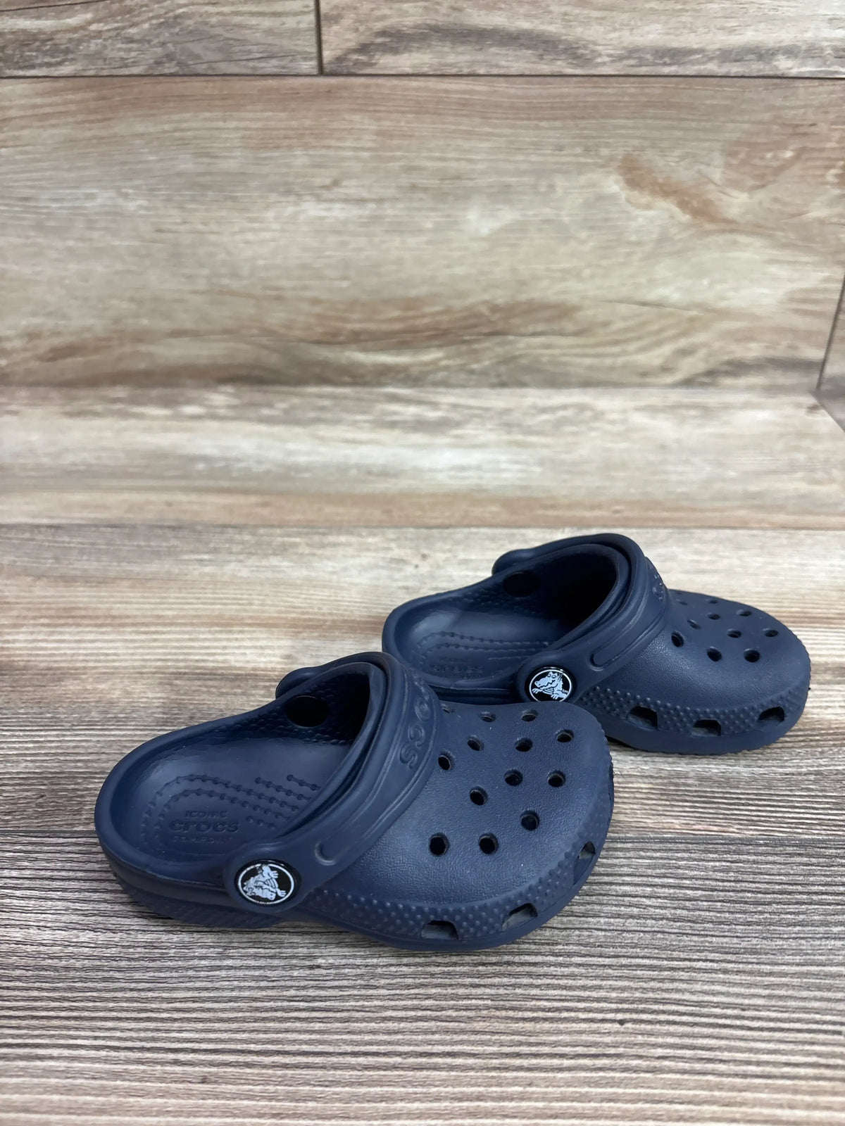 Angled view of navy Crocs Classic Clogs showcasing ventilation holes and heel straps on a wood-grain background