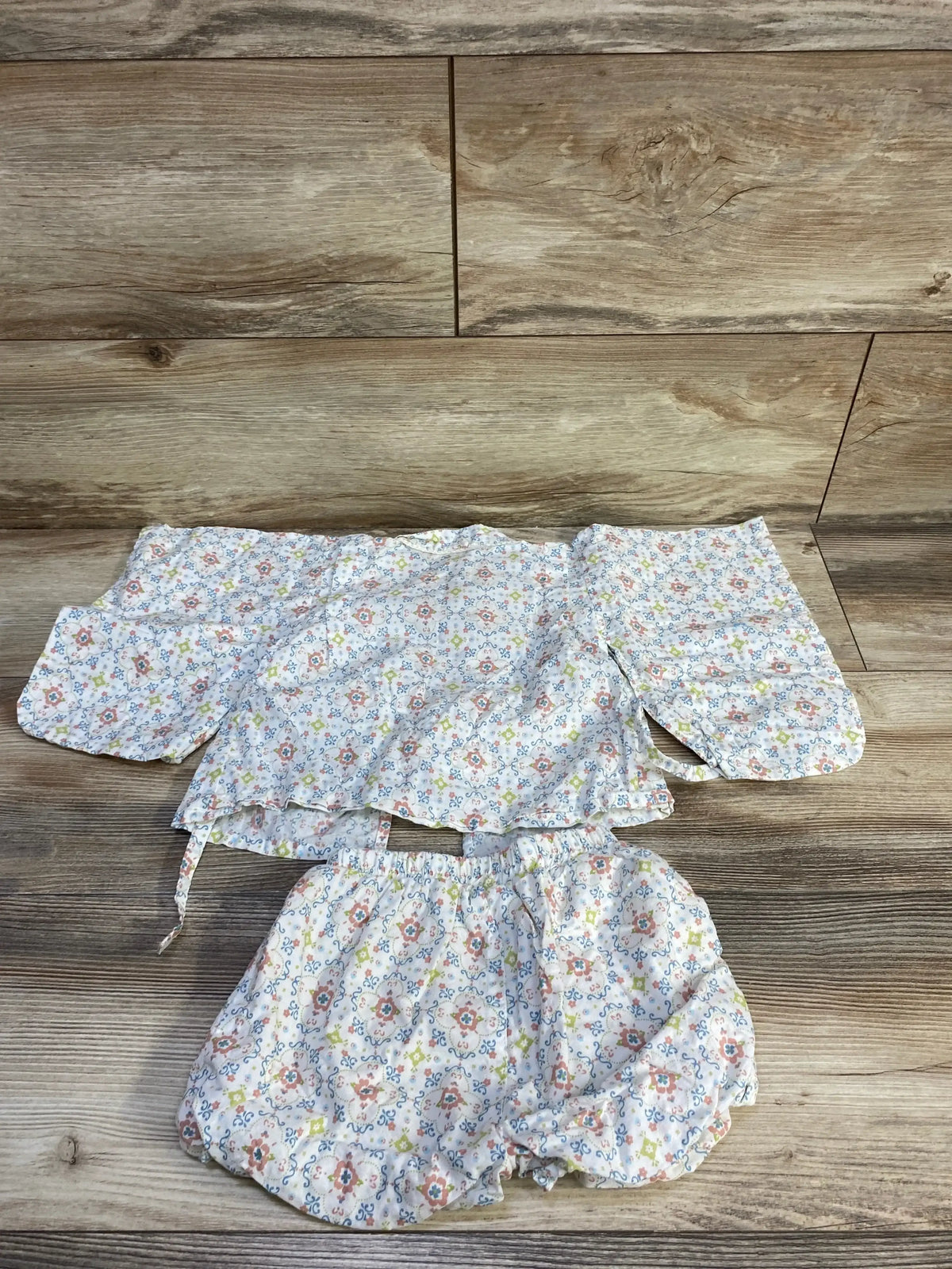 Flat lay of Tete a Tete floral wrap-style top and shorts set in white, showcasing an all-over floral print on a wood background