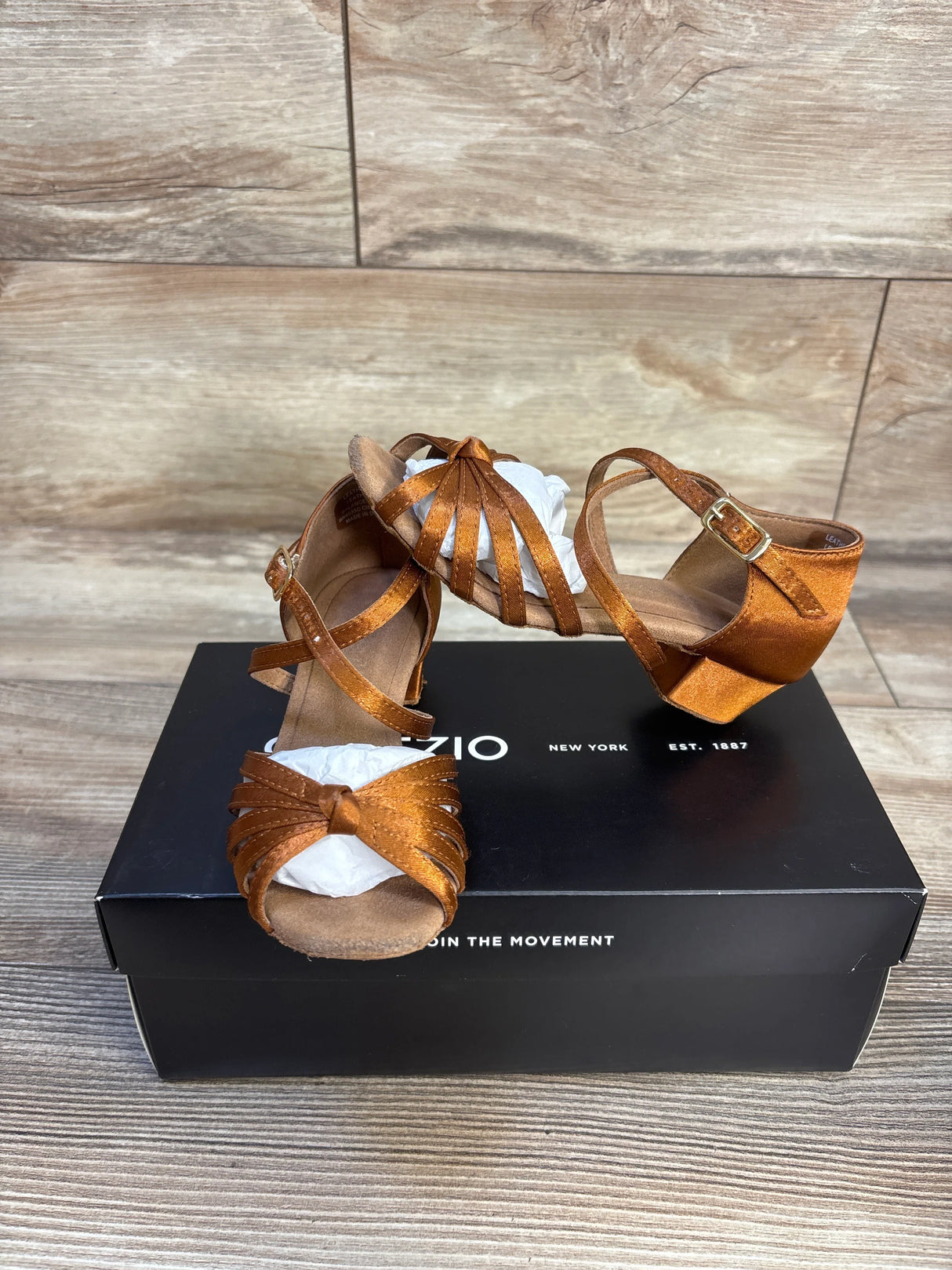 Three-quarter view of Capezio Valentina ballroom flats in cinnamon showing strappy toe, ankle buckle and heel on black box