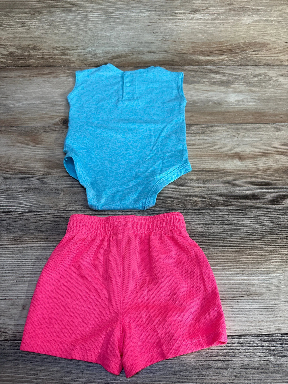 Front view of Under Armour two-piece bodysuit and shorts set showcasing a blue sleeveless bodysuit and bright pink shorts on wood flooring