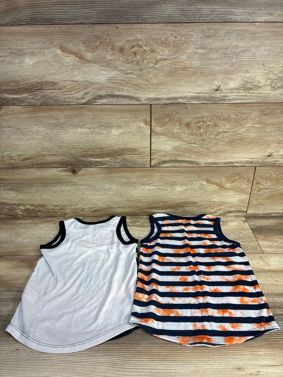 Flat lay of two toddler tank tops (size 4-5T): solid white with black trim and navy-and-white stripes with orange tie-dye, on wood background