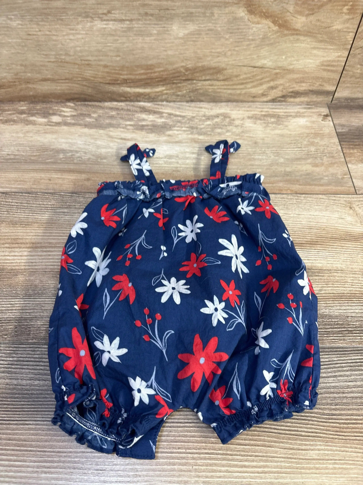 Flat lay of navy floral baby romper (3M) showing red and white flowers, elastic leg openings and shoulder tie straps on wood