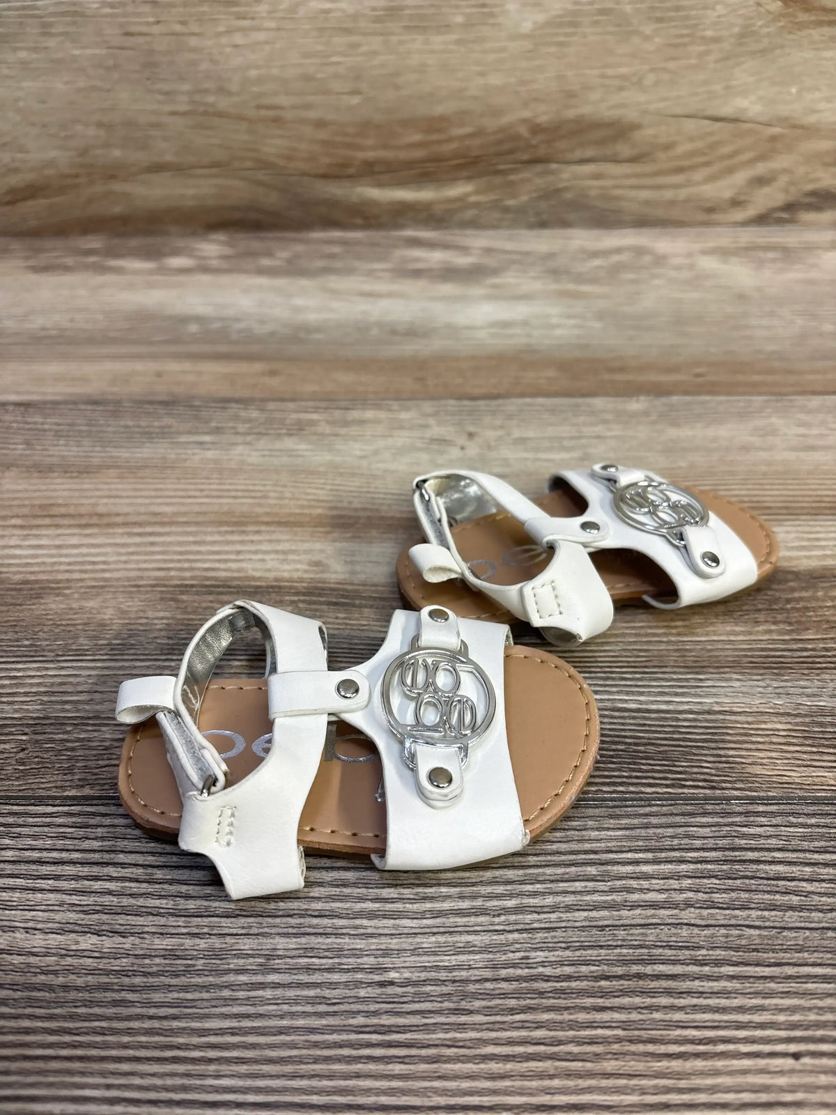 Overhead view of white Bebe sandals showcasing silver circular embellishments and tan soles on a wood-grain background