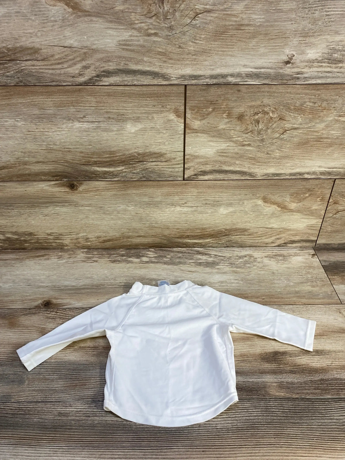 Back view of white iPlay baby rashguard, size 6 months, showing long sleeves and curved hem on wood background