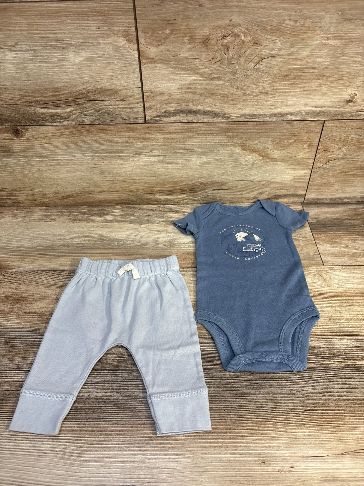 Baby blue onesie with adventure print and light gray pants on wood floor